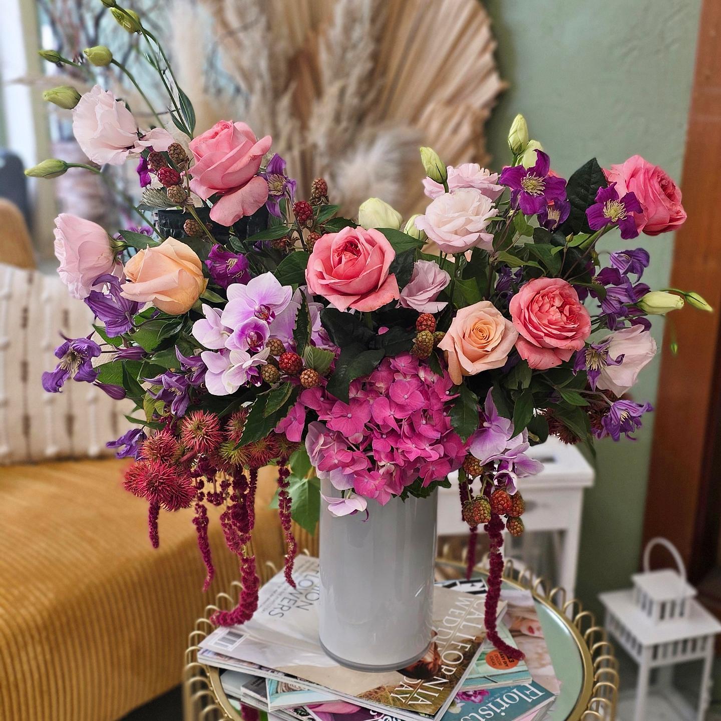 A large bouquet of pink, purple, and peach flowers in a white vase on a table, with a yellow sofa and green wall in background.