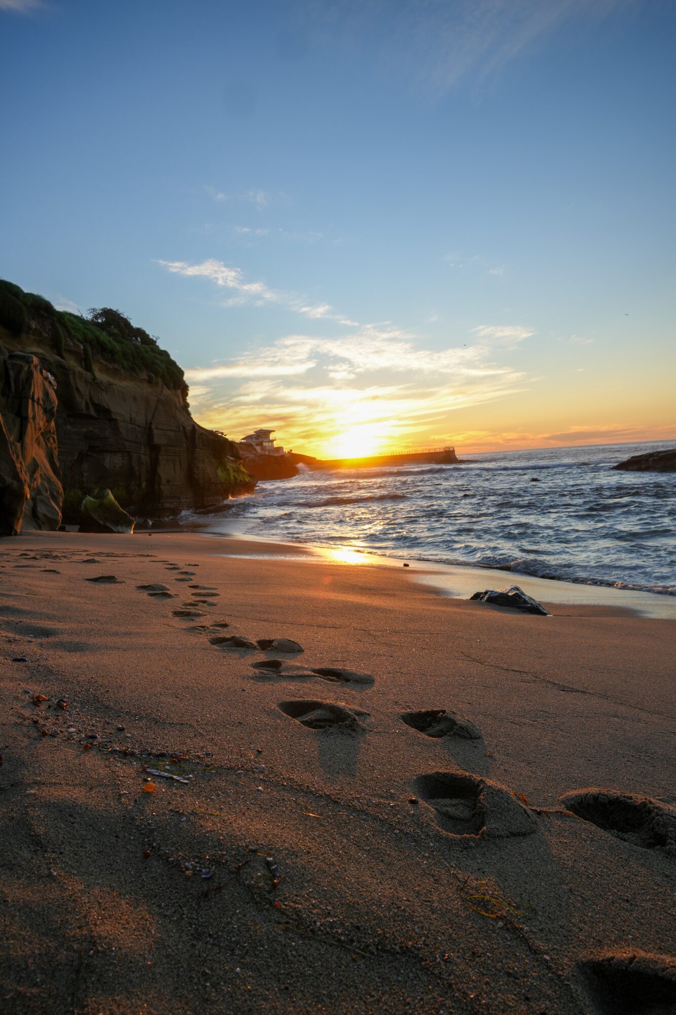 Sunset over a beach with footprints in the sand, cliffs on the left, and the ocean on the right.