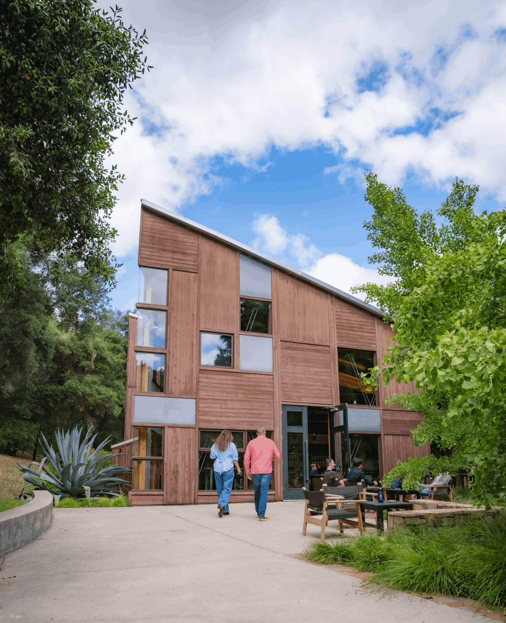 Three people walk toward a modern wooden building with large windows, surrounded by trees and outdoor seating, under a partly cloudy sky.
