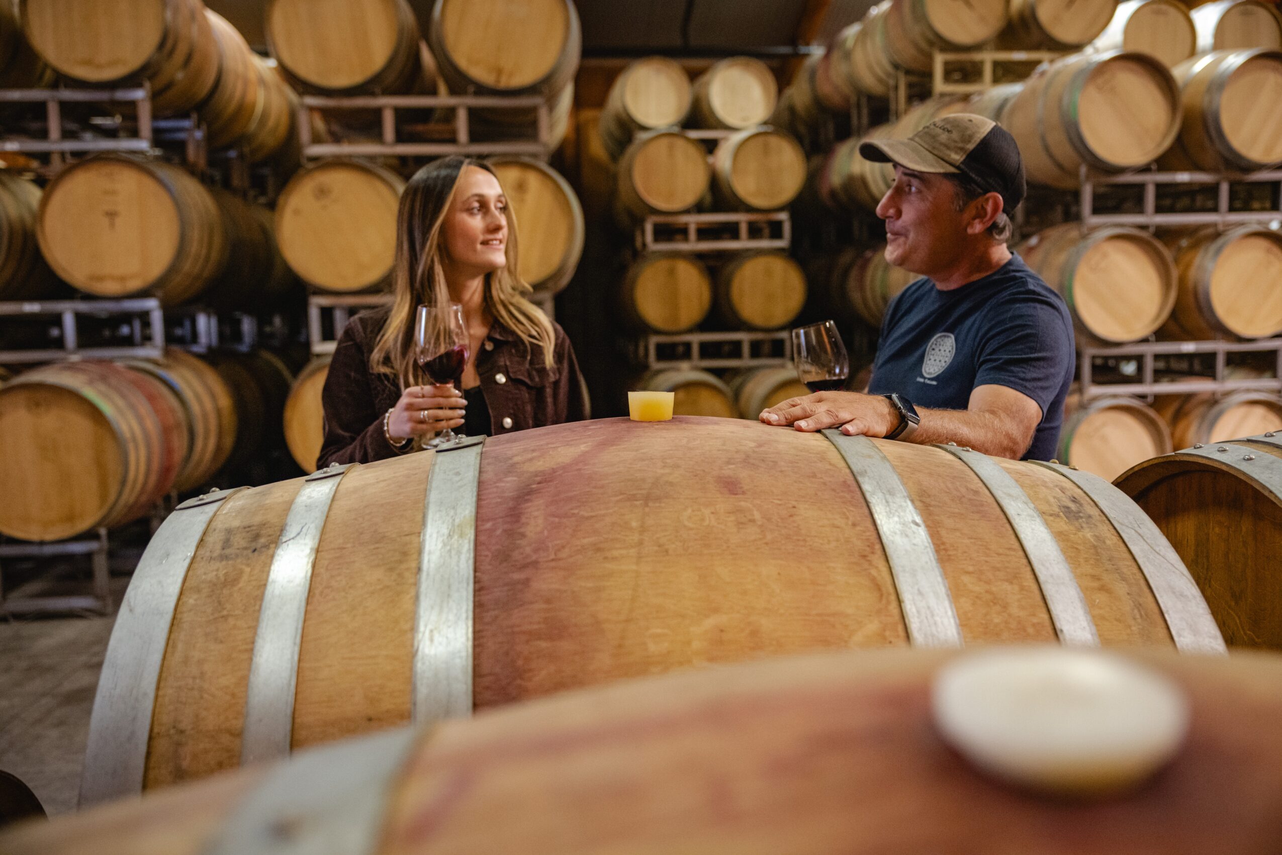 Two people in a wine cellar with barrels, one holding a glass and the other smiling, engaged in conversation.