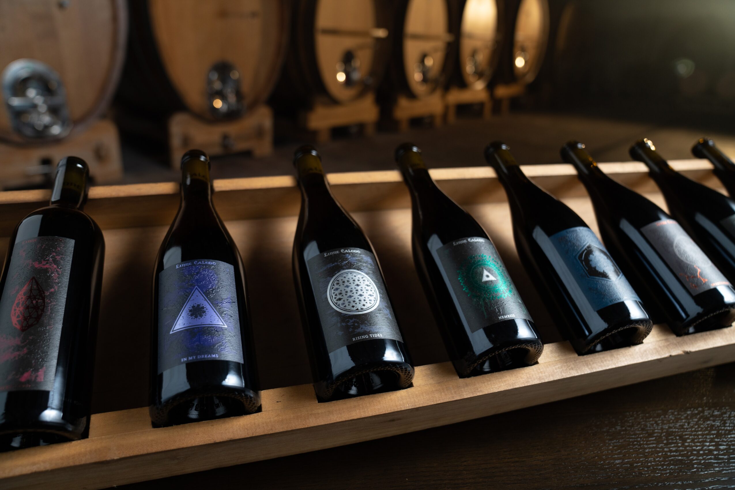 Six bottles of wine on a wooden rack with barrels in the background, viewed from the side.