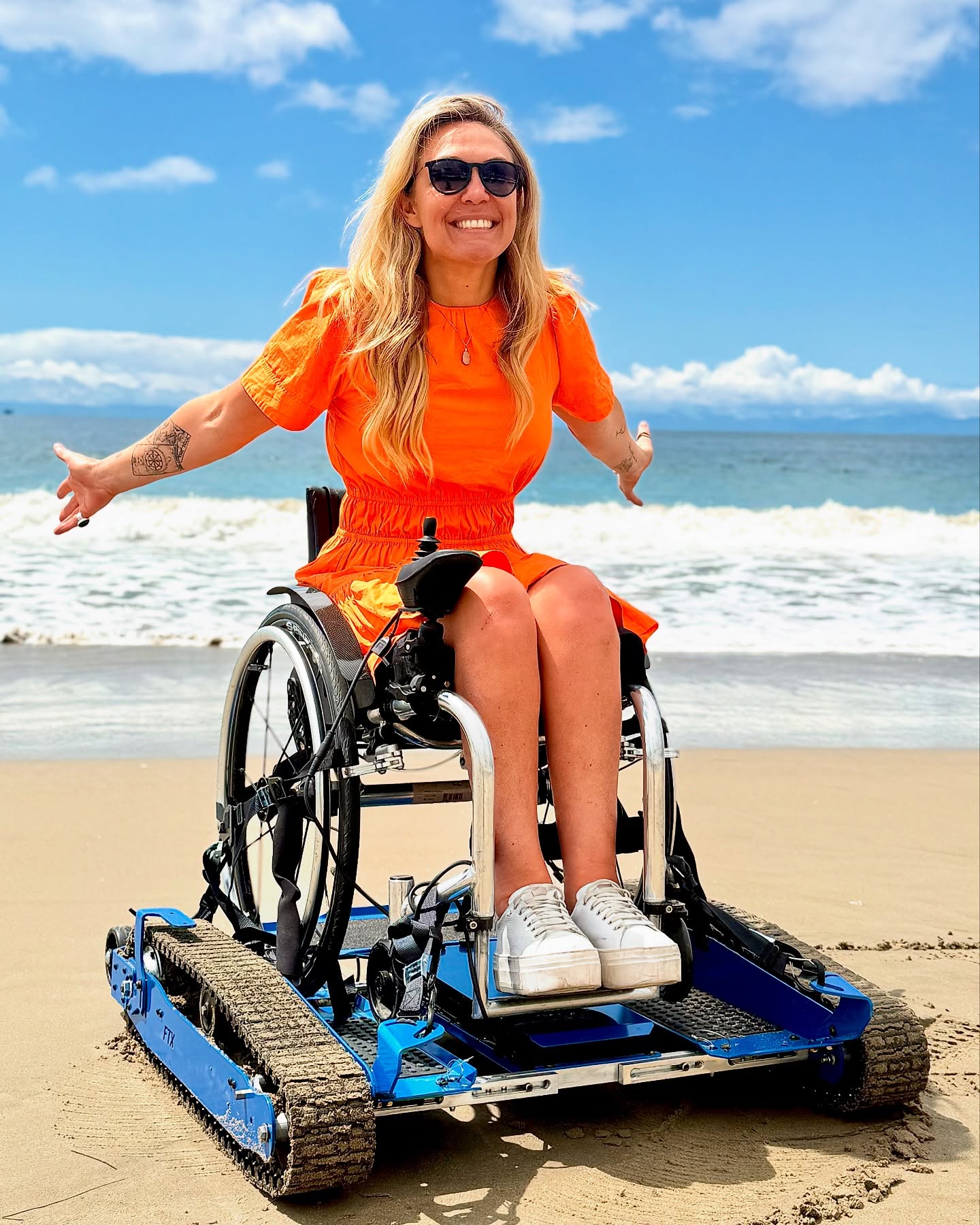 Woman in orange dress in a wheelchair on beach, smiling, with ocean and sky in background.