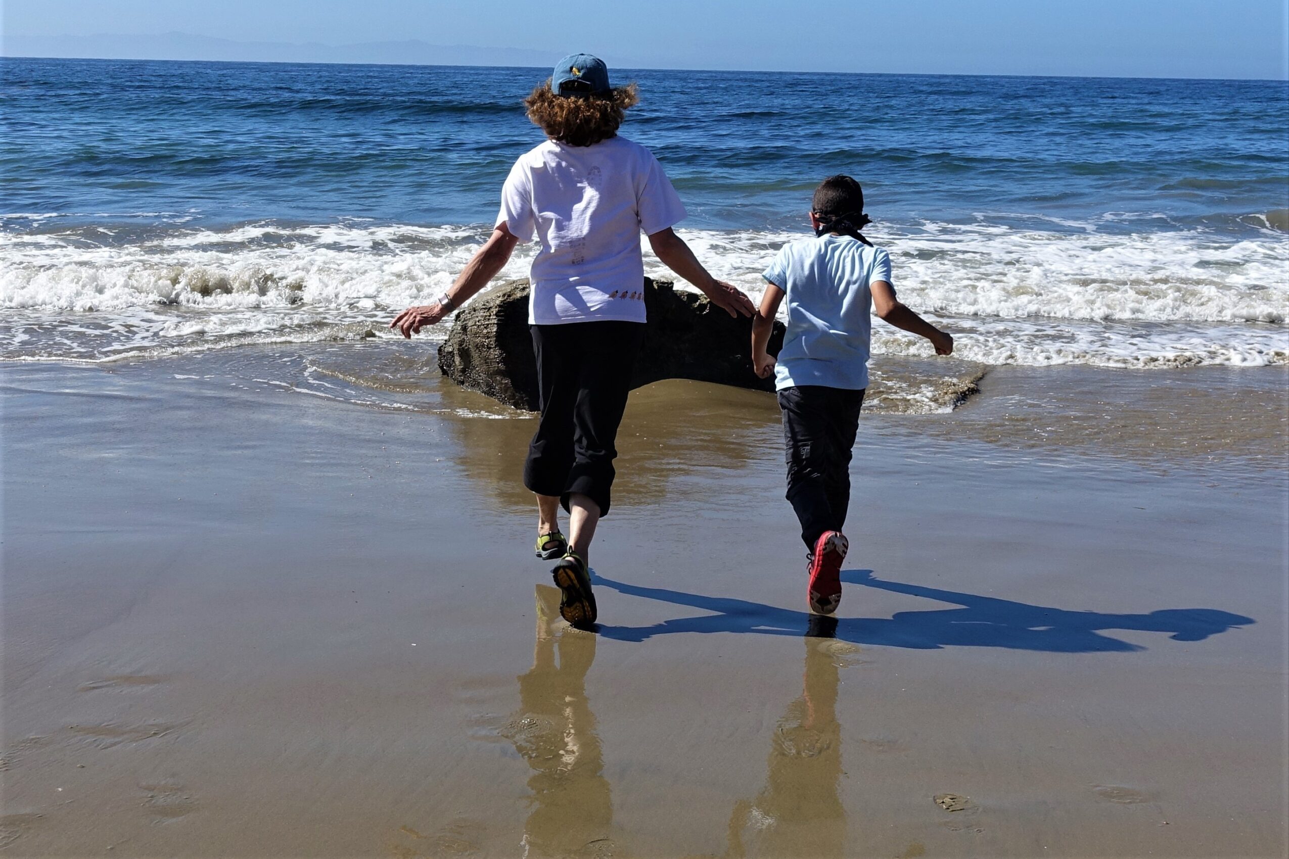 Adult and child holding hands walking into the ocean on a sandy beach, waves in the background, sunny day.