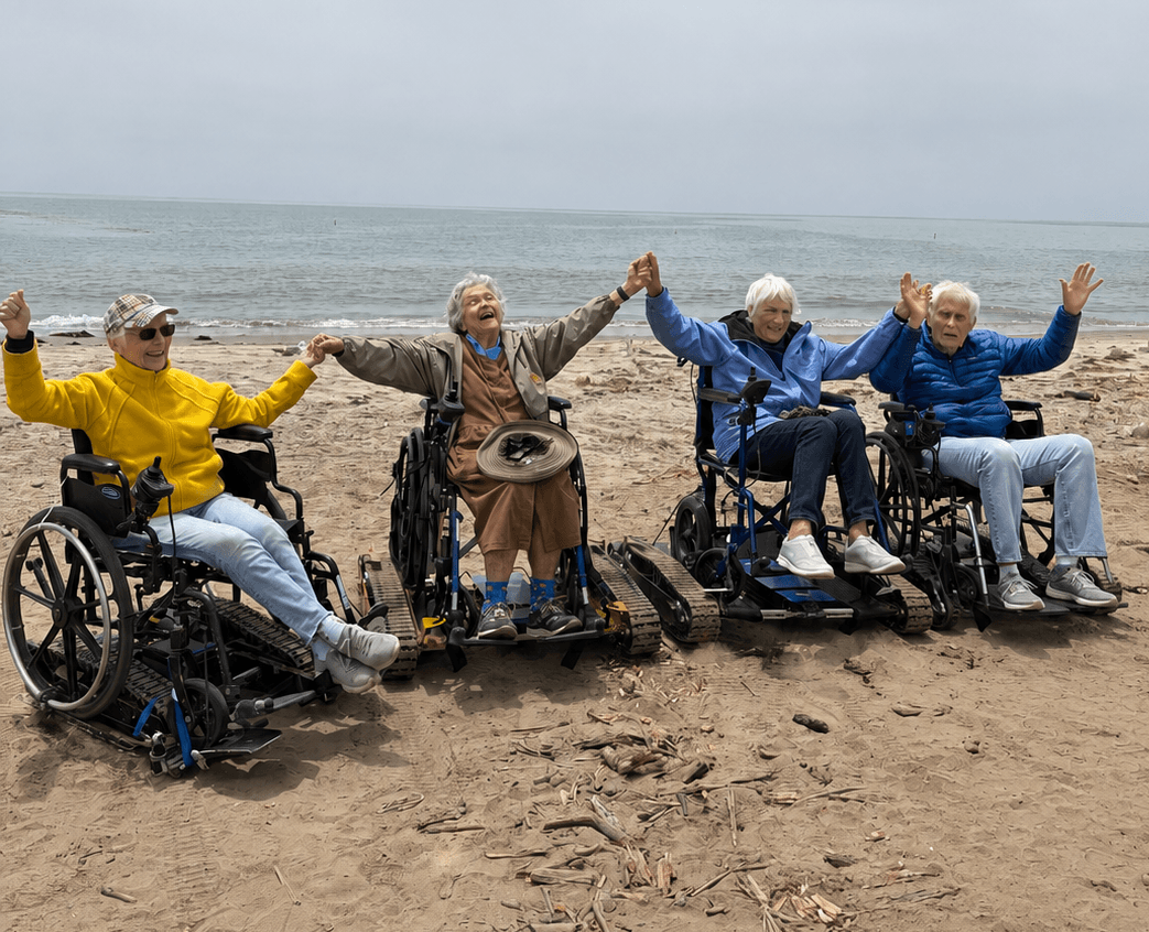 Four elderly people in wheelchairs on a beach, raising their hands and smiling, with the ocean in the background.