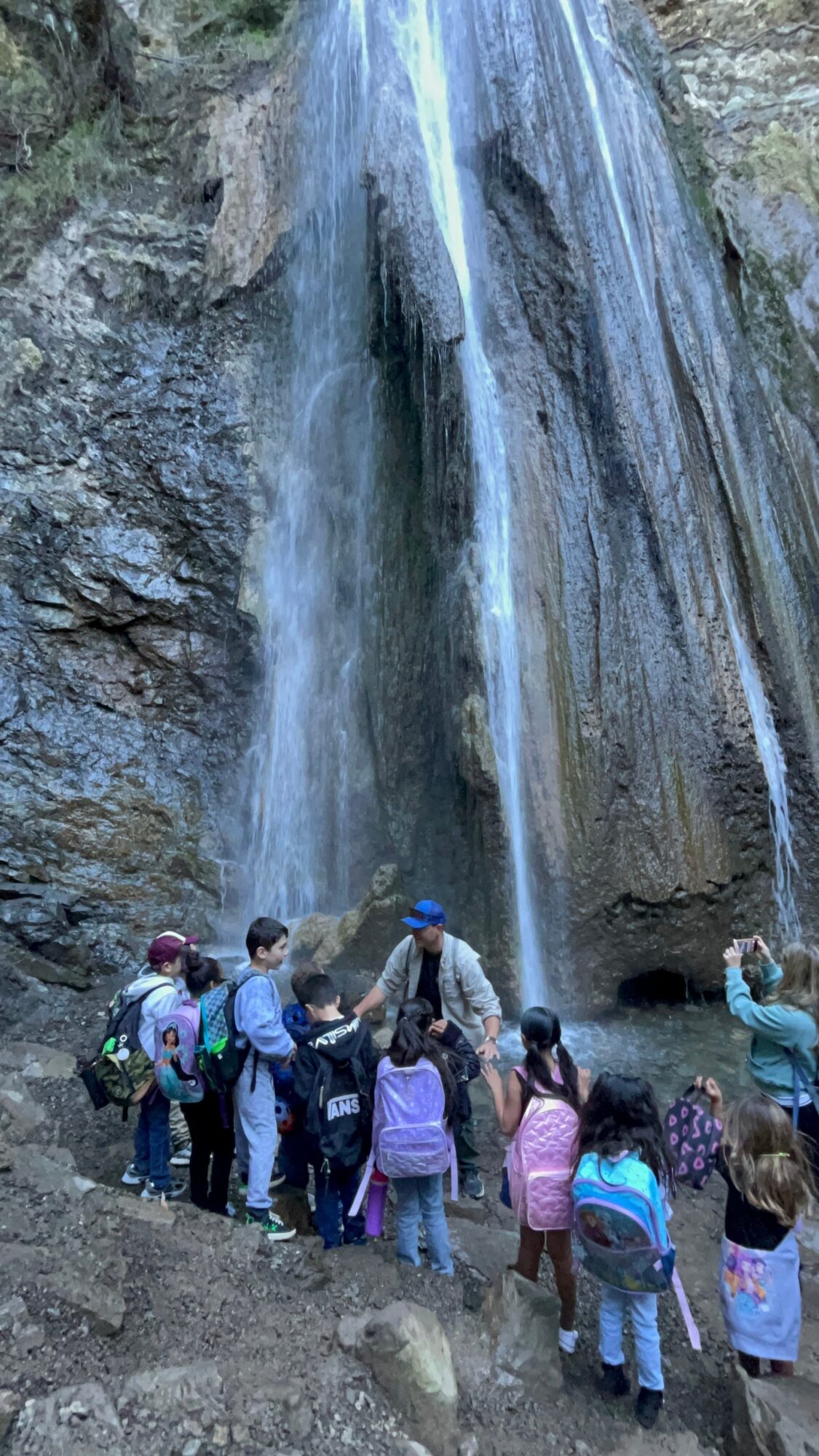 Group of children and adults standing near a waterfall on rocky terrain, some taking photos, wearing backpacks and colorful clothing.