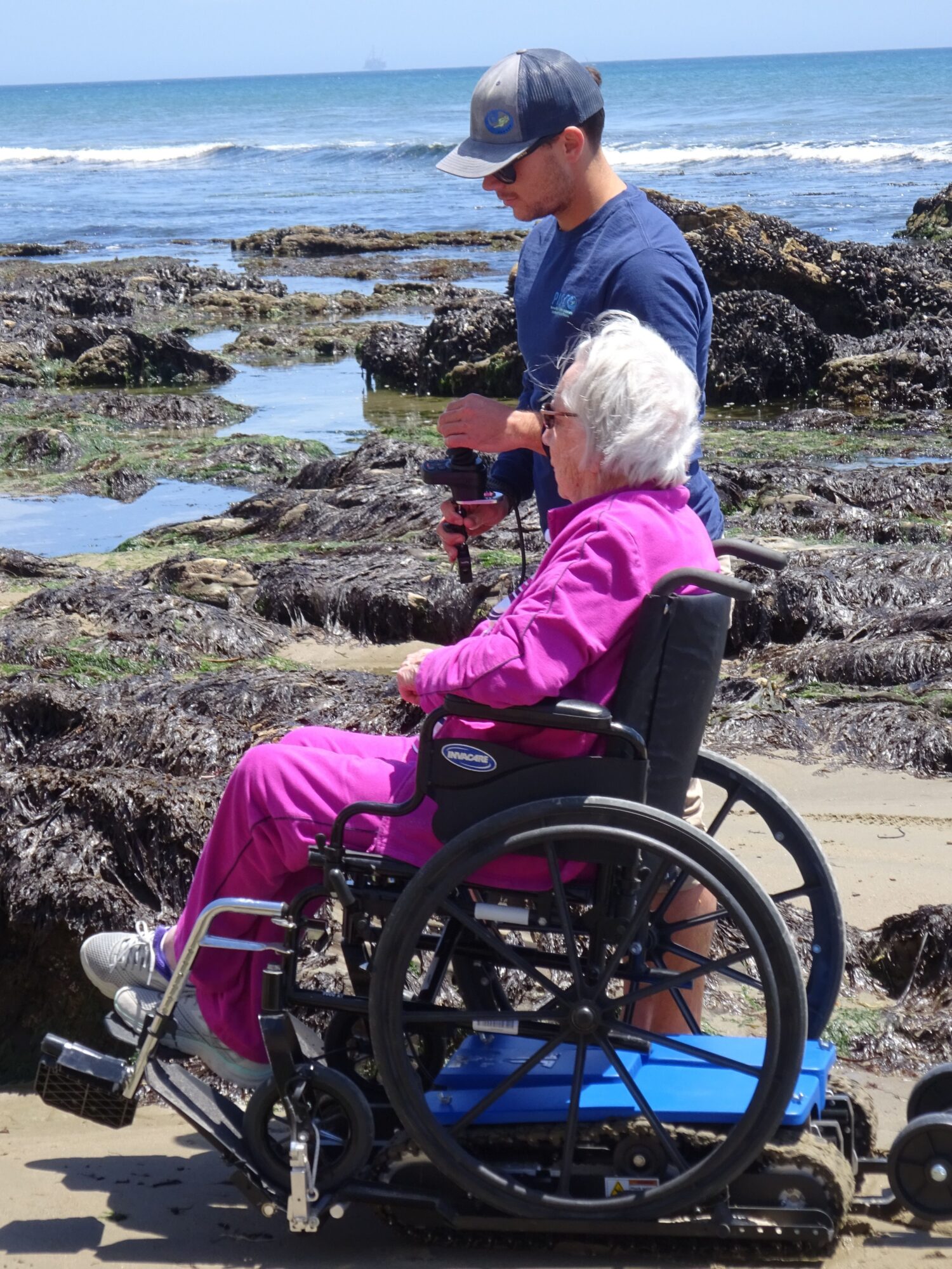 Two people on a rocky shoreline, one in a wheelchair, with ocean waves in the background.