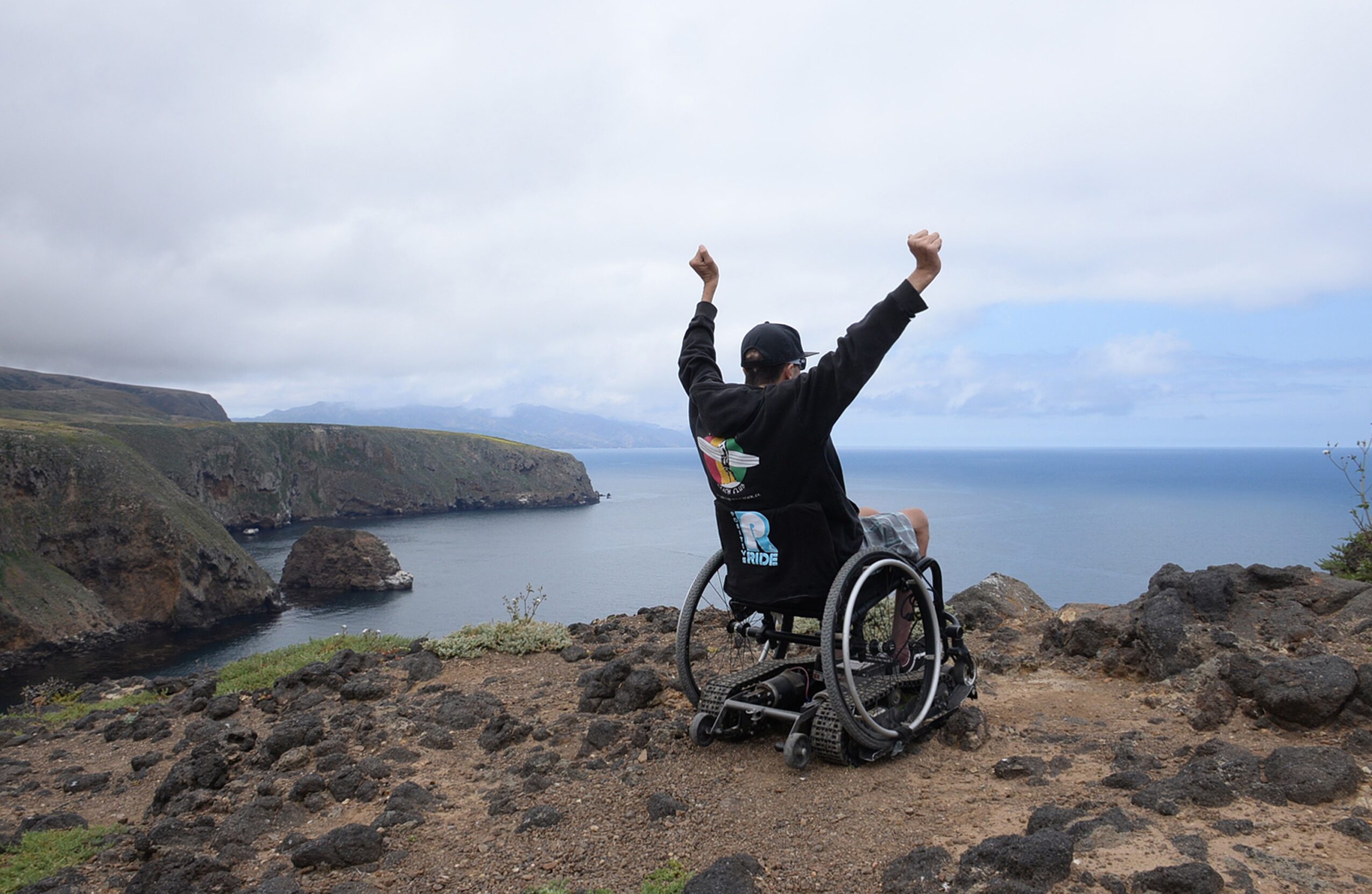 Person in wheelchair with arms raised, overlooking a coastal landscape with cliffs and ocean, cloudy sky.