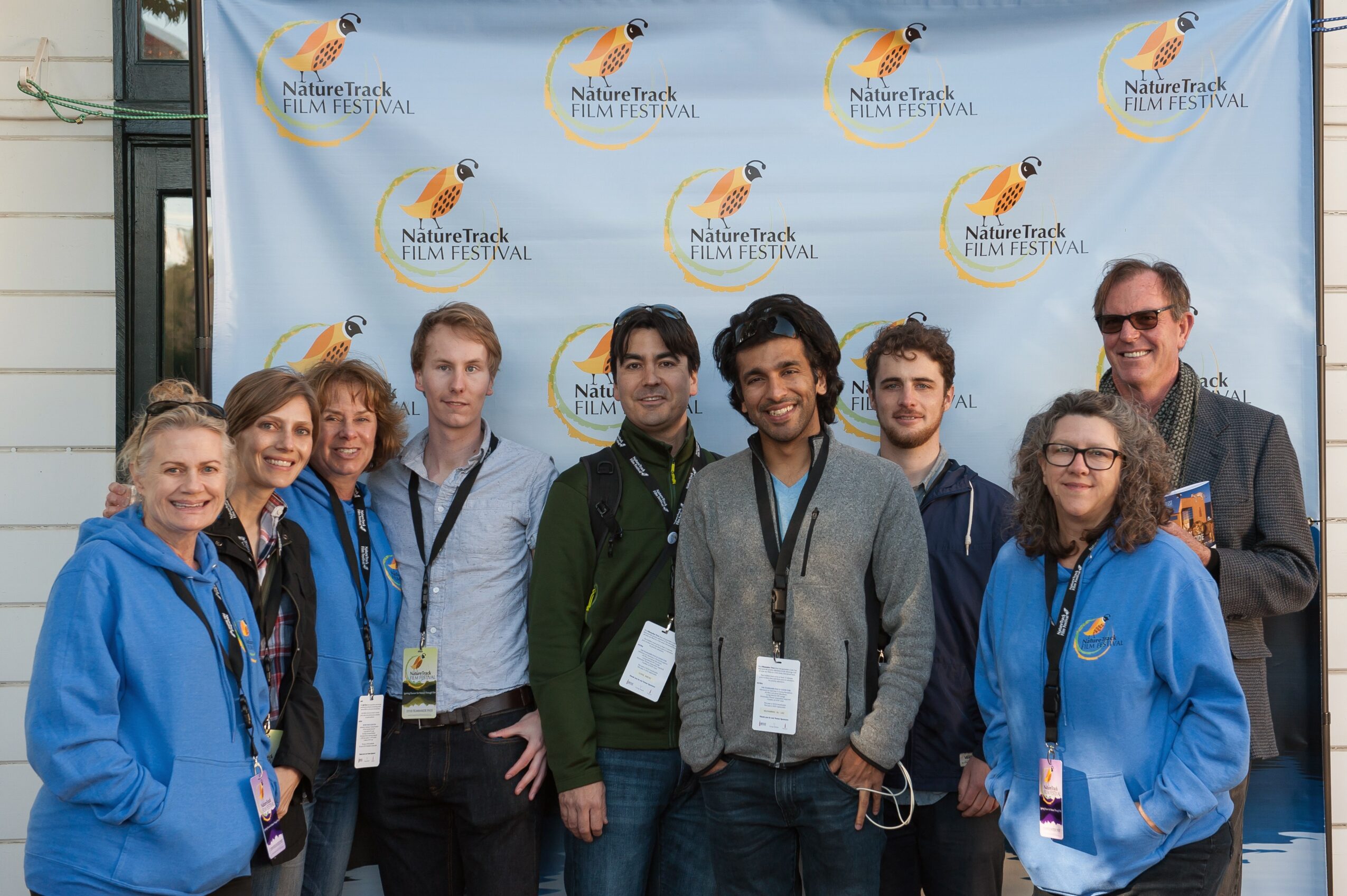 Group of nine people standing in front of a blue backdrop with festival logos, smiling for the photo.