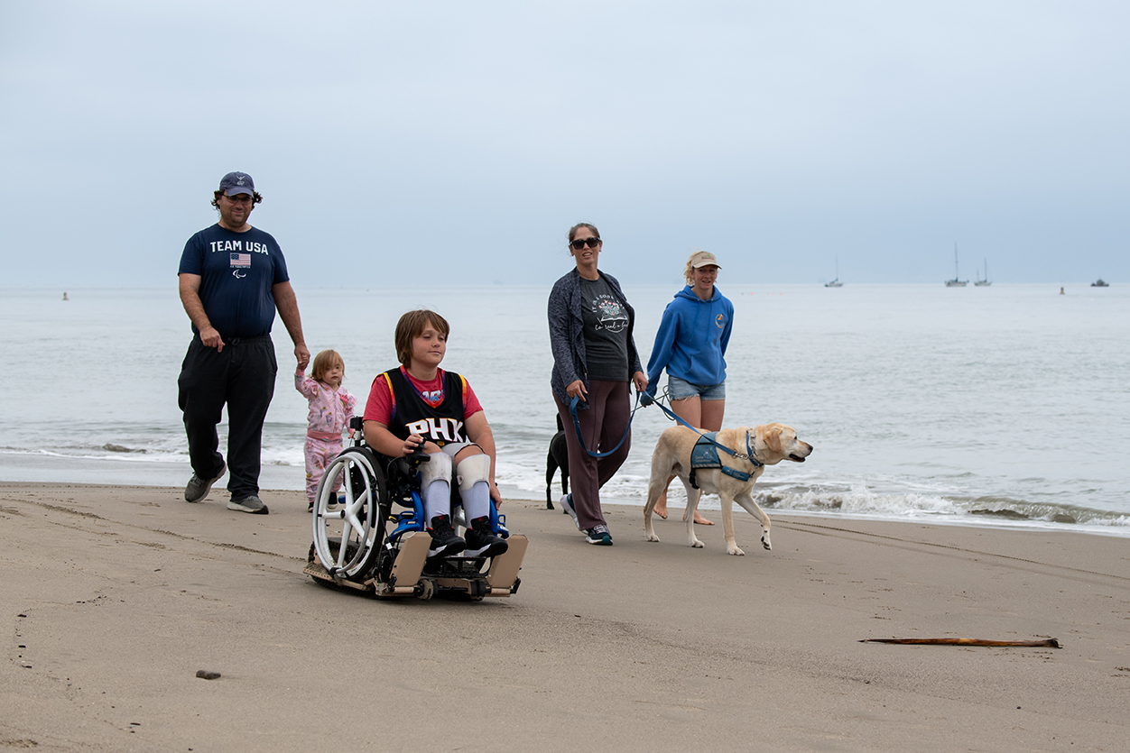 Four people and a dog walk along the beach with the ocean in the background, overcast sky, and boats in the distance.