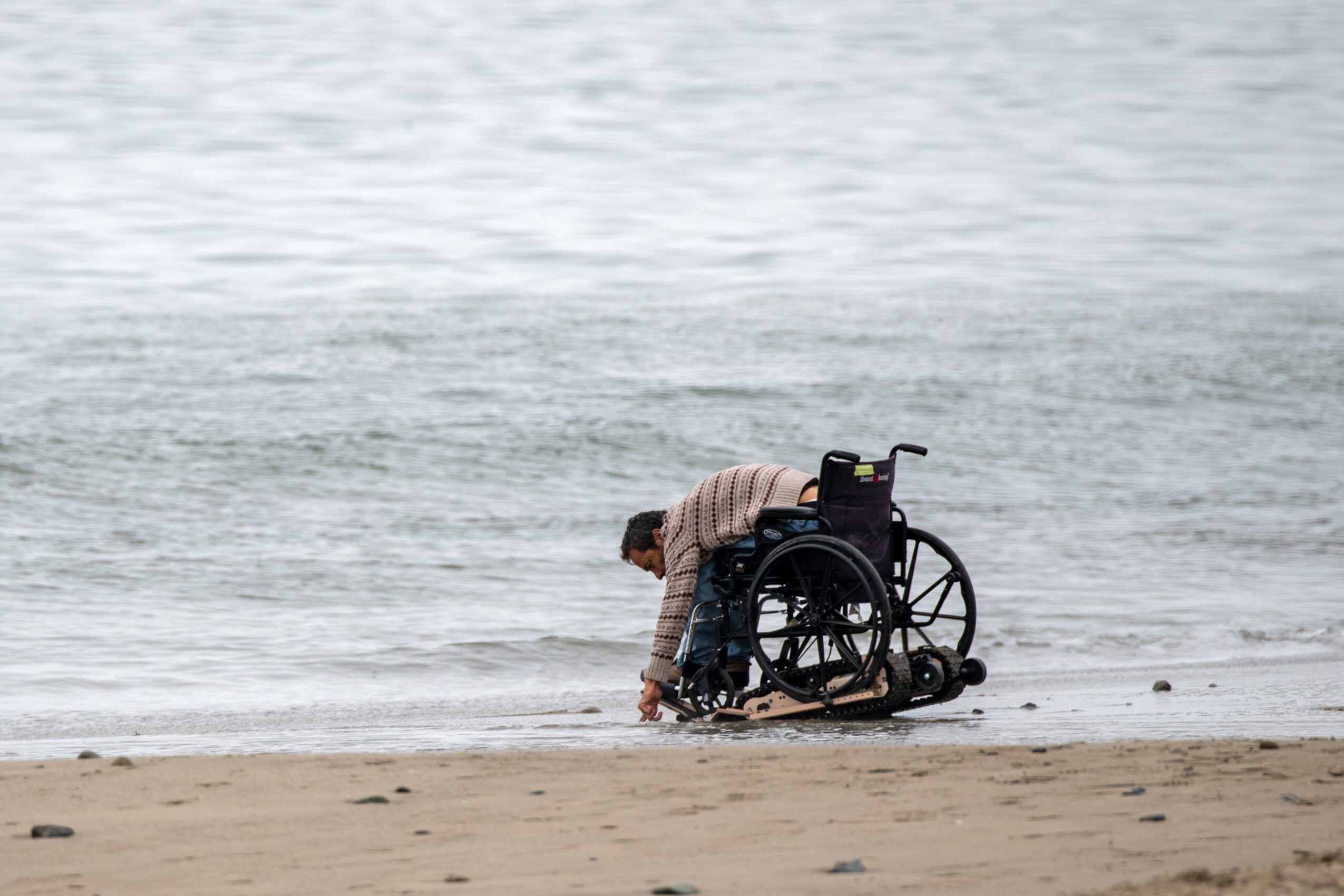 Person in a wheelchair on the beach, leaning forward near the water's edge, with waves in the background.