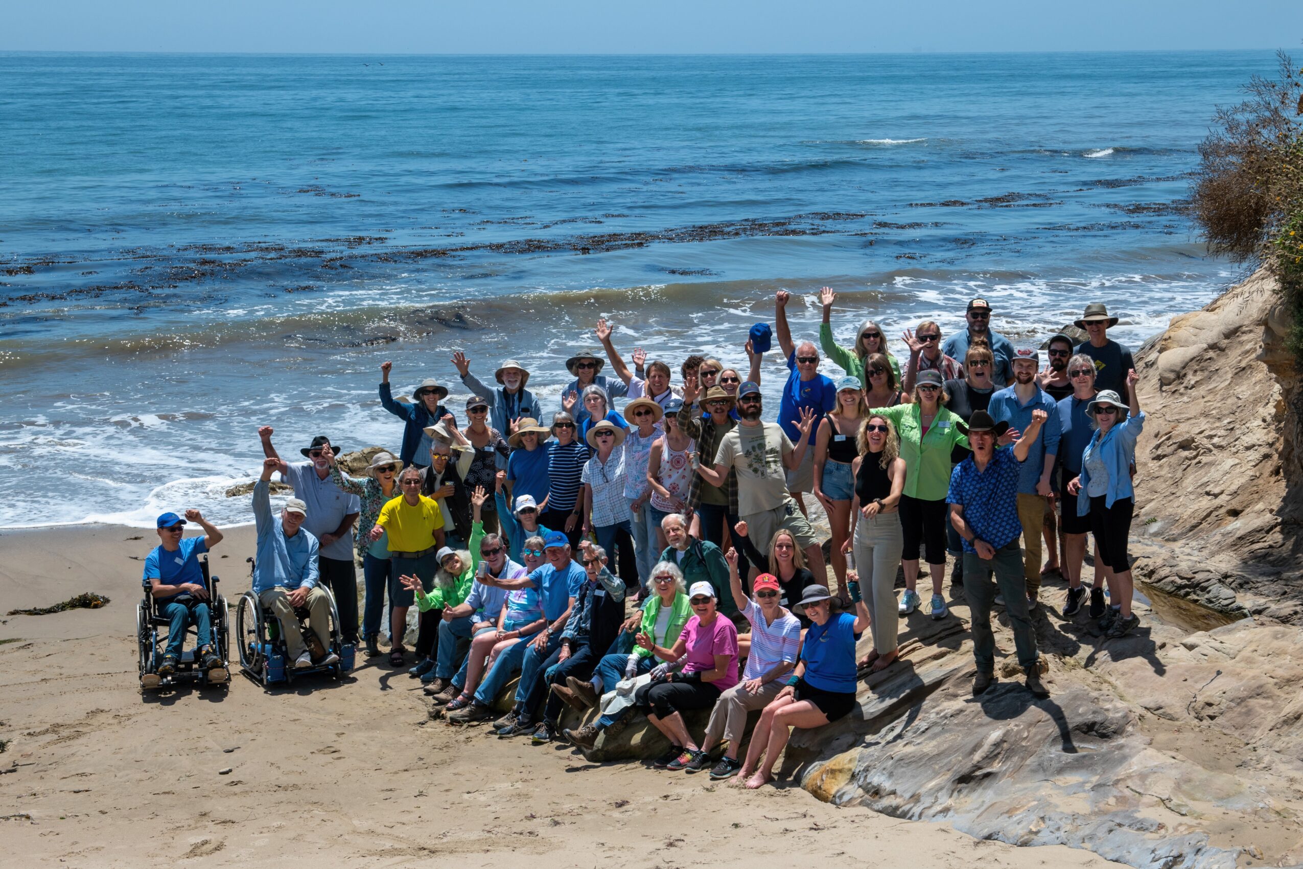 Group of people on a beach, some in wheelchairs, waving at the camera with ocean and sky in background.