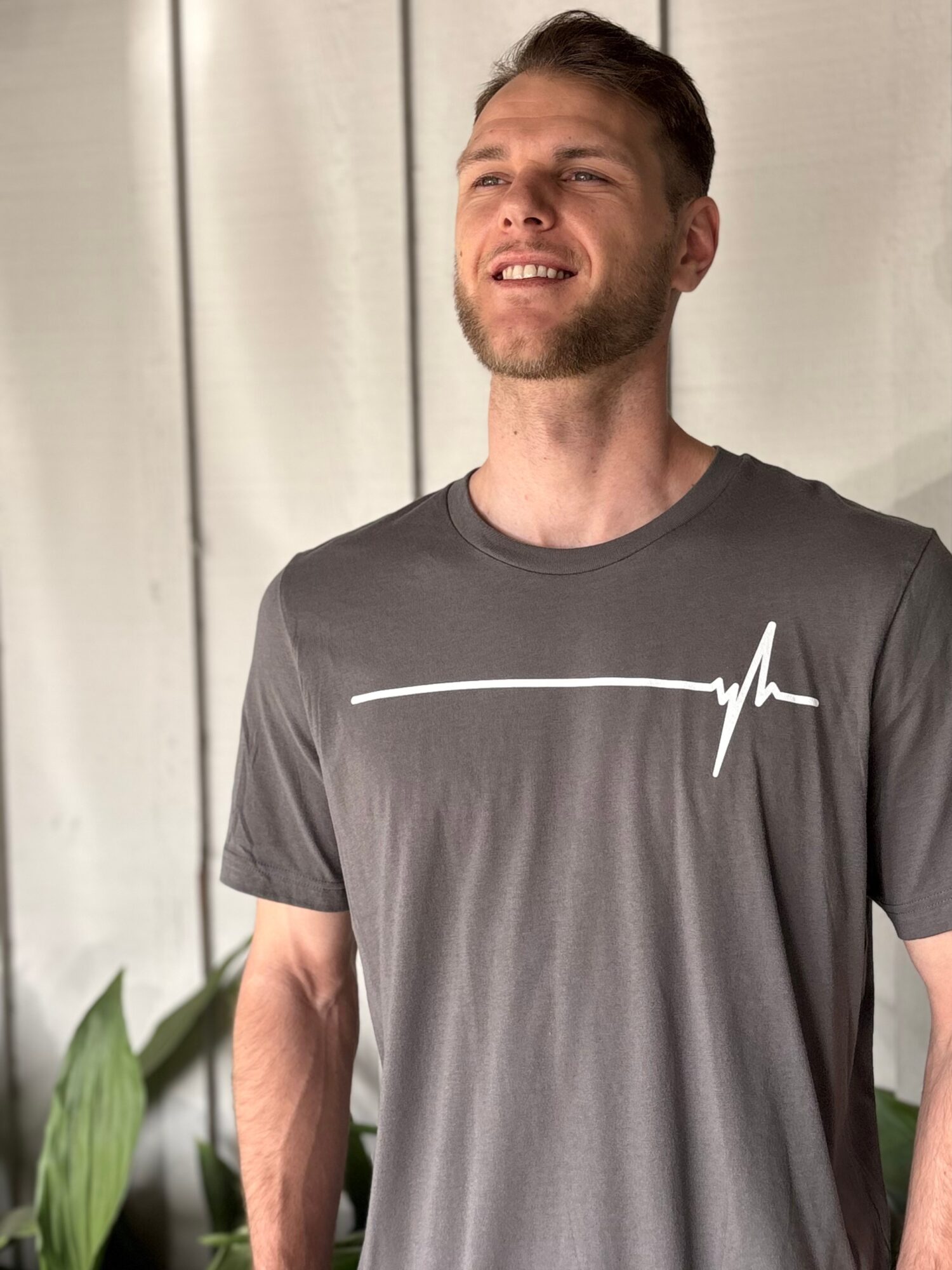 Young man with short hair and beard smiling outdoors, wearing a gray T-shirt with a white line and heartbeat symbol.
