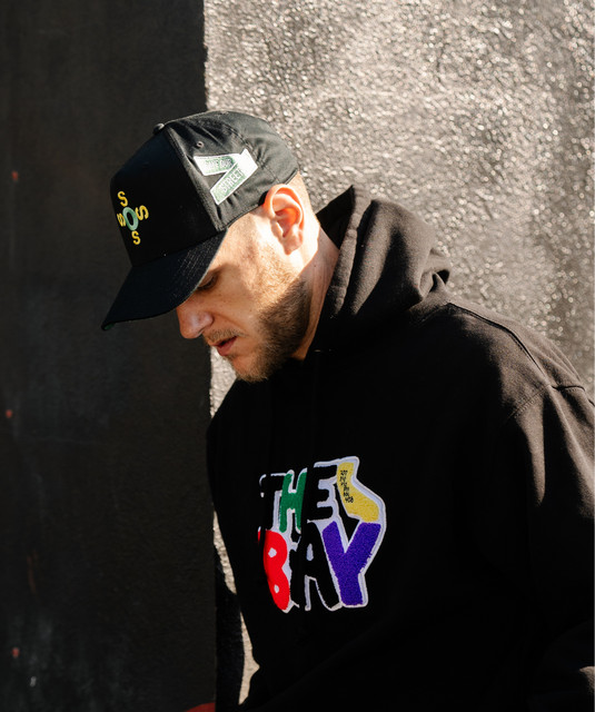 Young man wearing a black cap and hoodie, looking downward, standing against a textured wall.