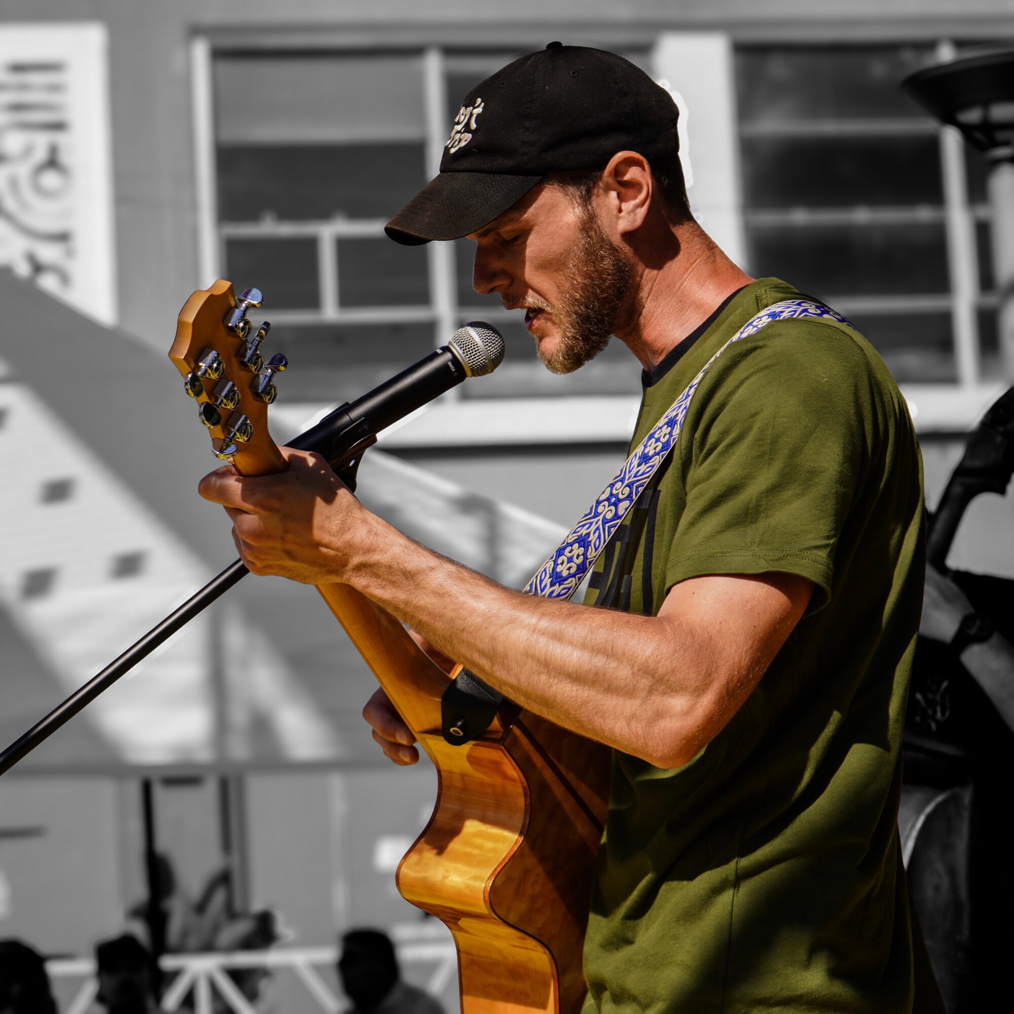 Man playing guitar and singing into microphone outdoors, wearing a black cap and green shirt.