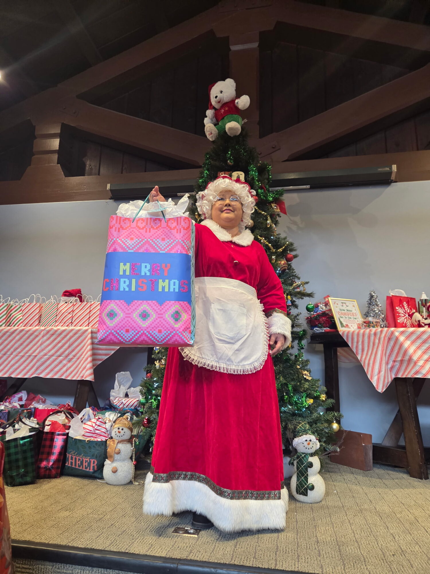 Person dressed as Santa Claus standing in front of a decorated Christmas tree, holding a gift bag, with wrapped presents nearby.