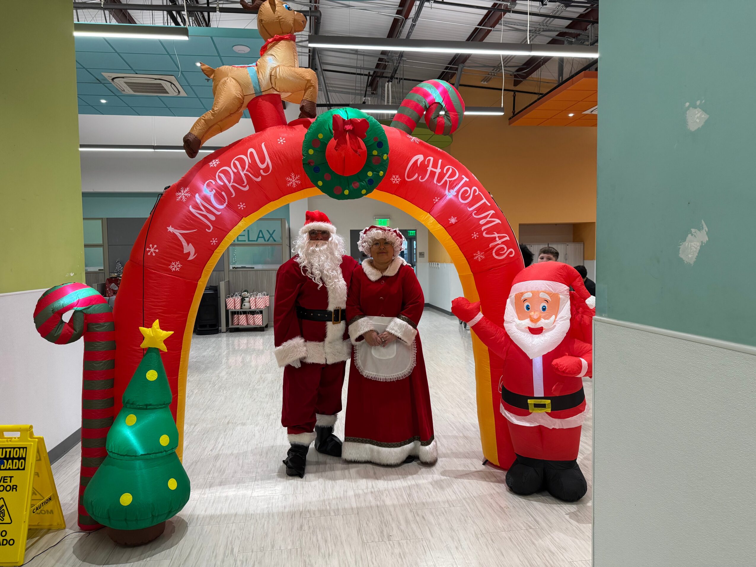 Two people dressed as Santa Claus and a woman in holiday costumes stand under a Christmas arch with festive decorations.