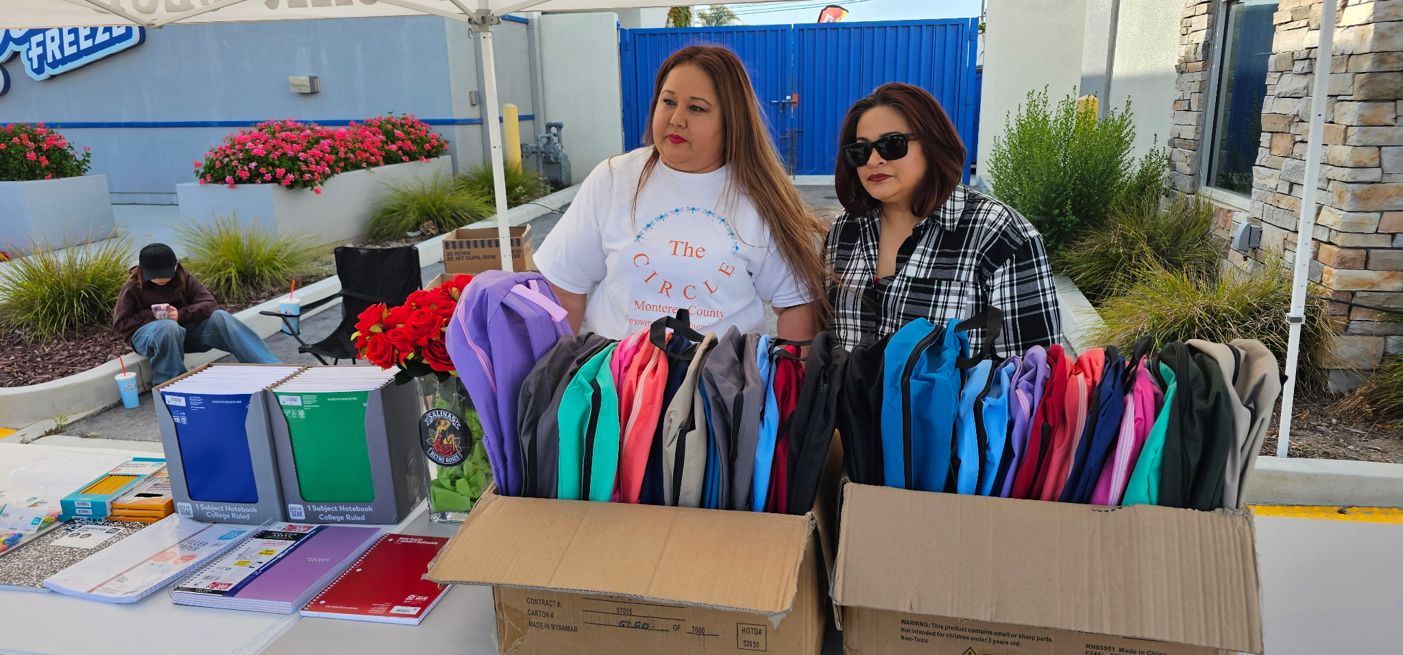 Two women stand behind a table with colorful backpacks and folders, outdoors near plants and a building.