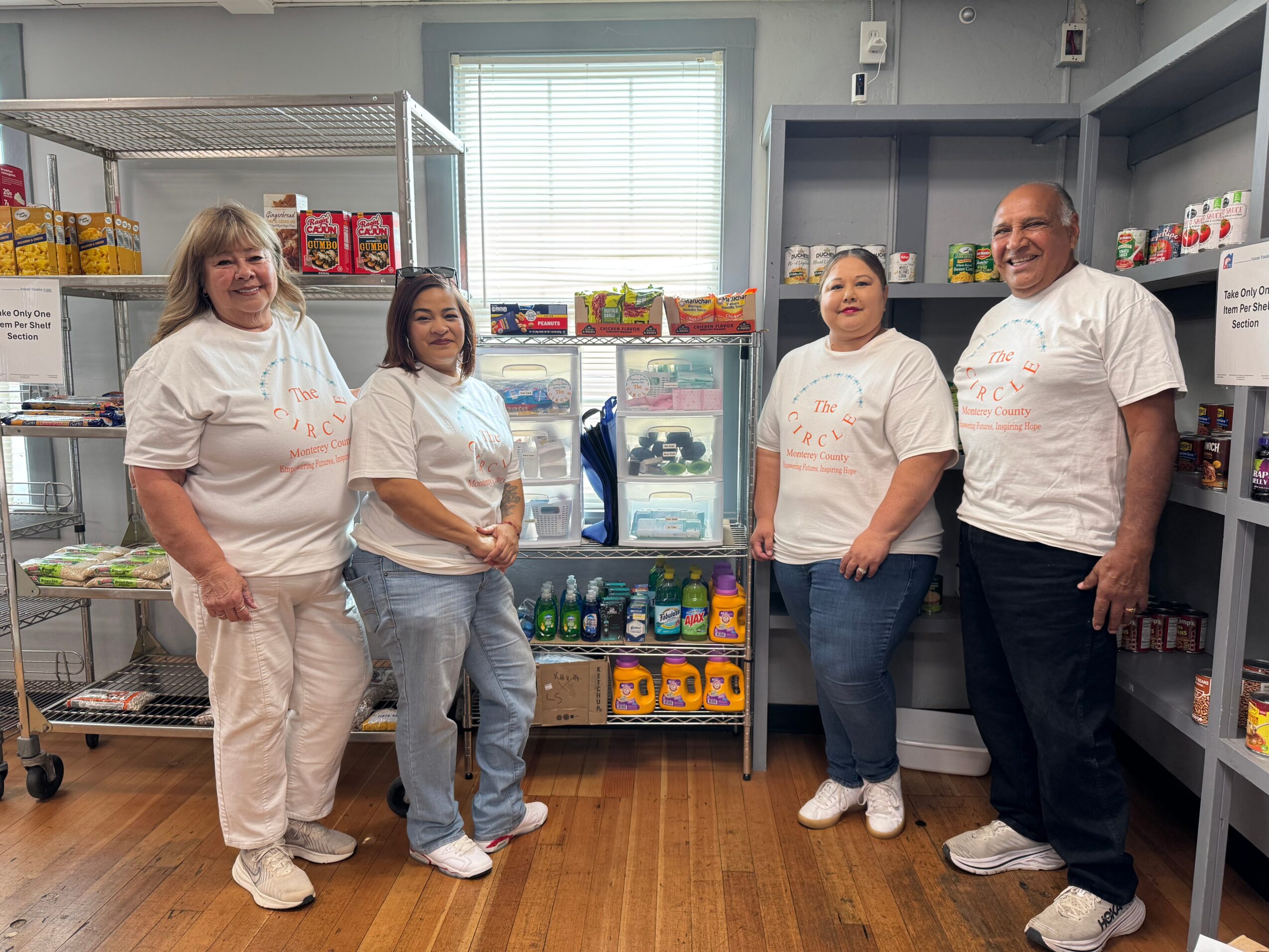 Four people standing in a store aisle with shelves and a refrigerator behind them, smiling and wearing white shirts.