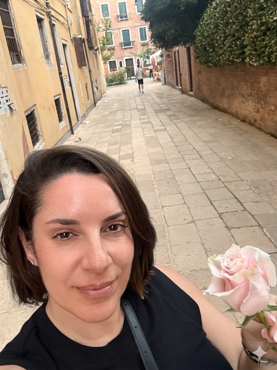Woman with shoulder-length dark hair taking a selfie on a narrow street with buildings and trees in the background.