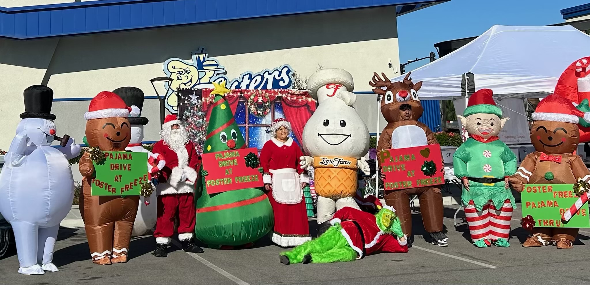 Group of people in Christmas costumes and mascots outdoors, with a building and tents in the background.