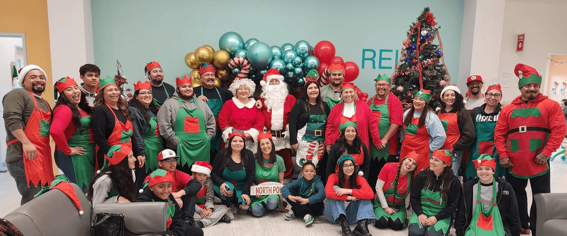 Group of people dressed as Santa and elves celebrating Christmas with a decorated tree and balloons.