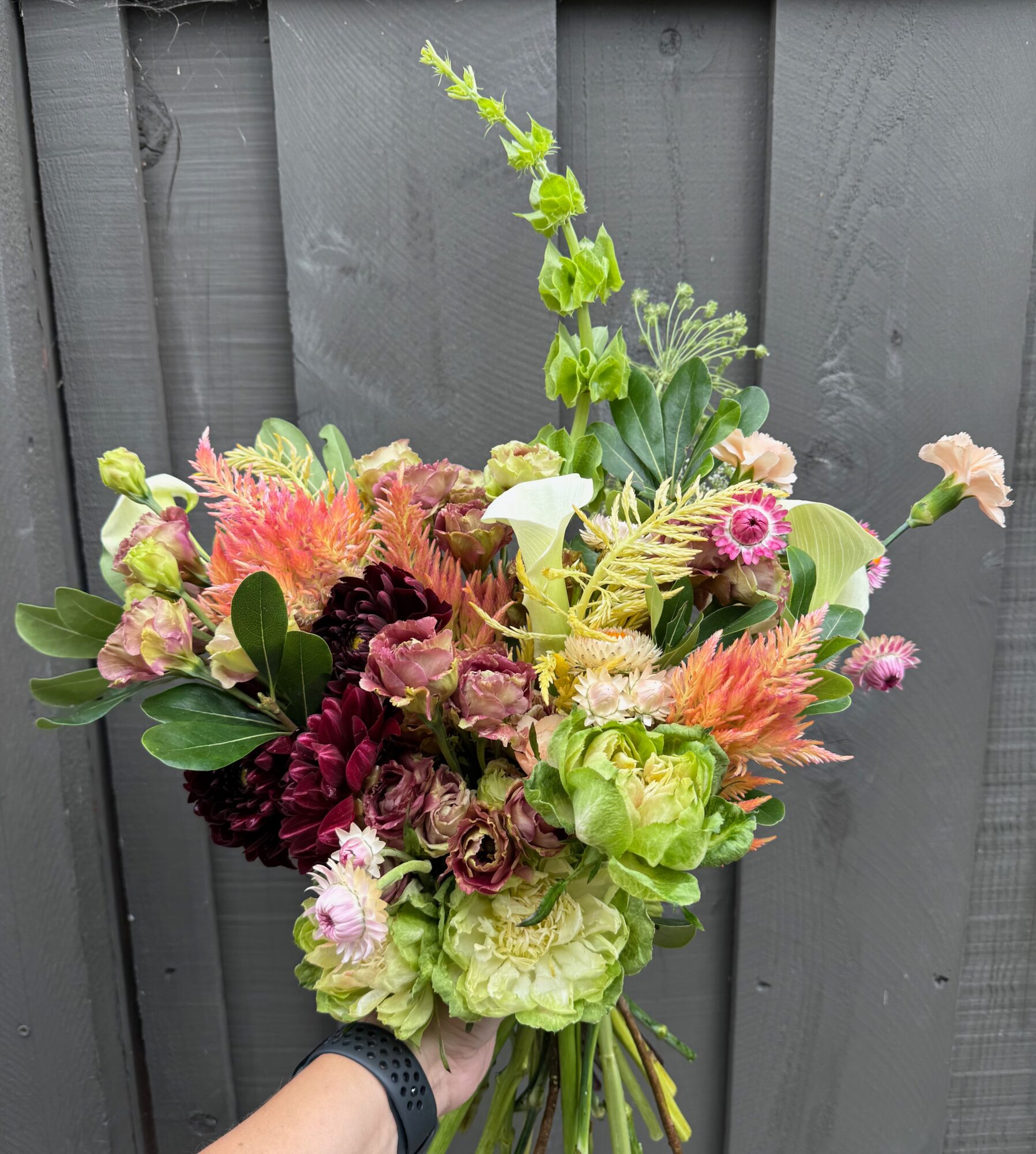 Hand holding a colorful bouquet of flowers with pink, green, and peach blooms against a gray wooden background.