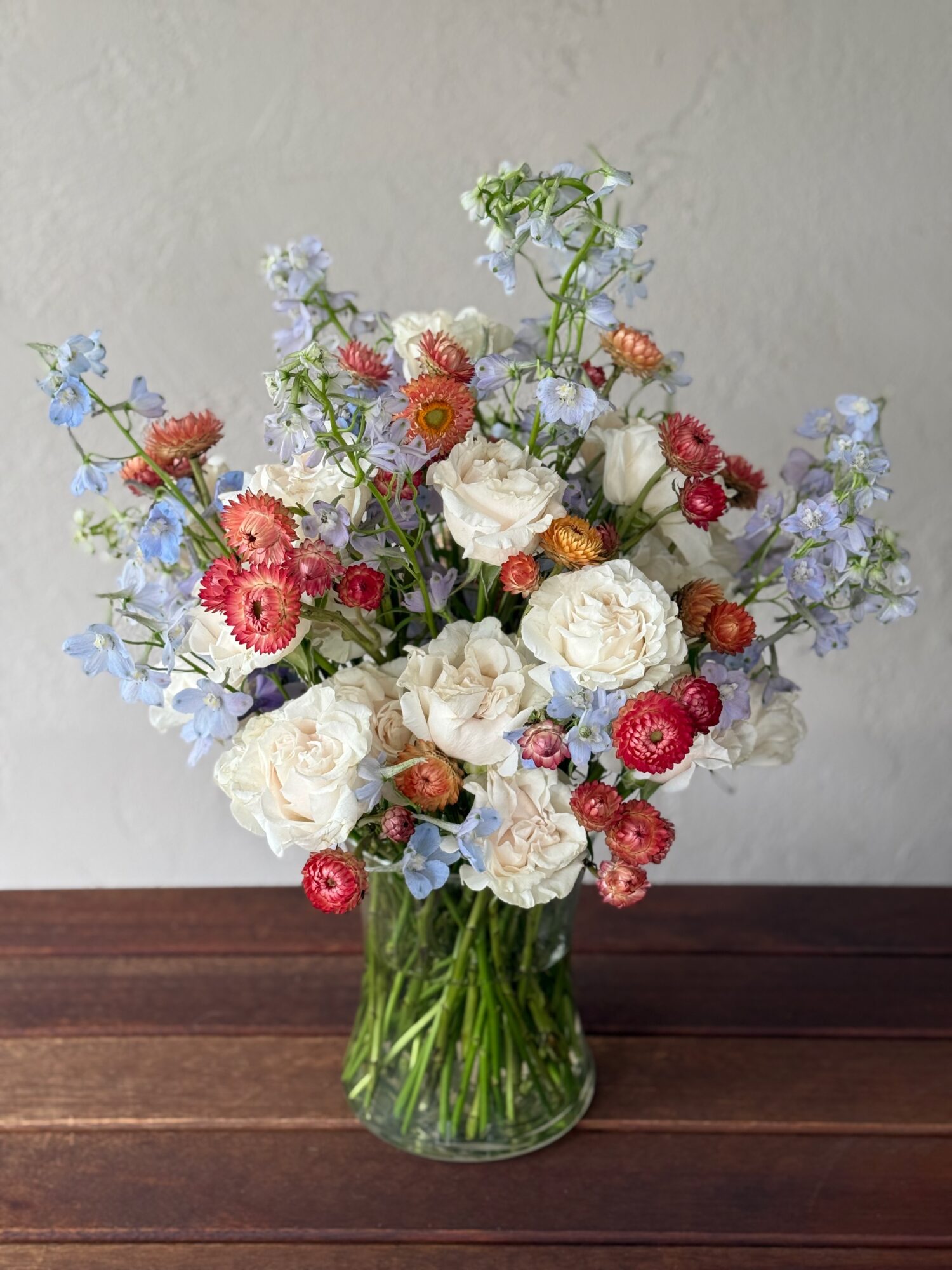 A colorful flower bouquet in a glass vase on a wooden surface, with white, red, pink, and blue flowers.