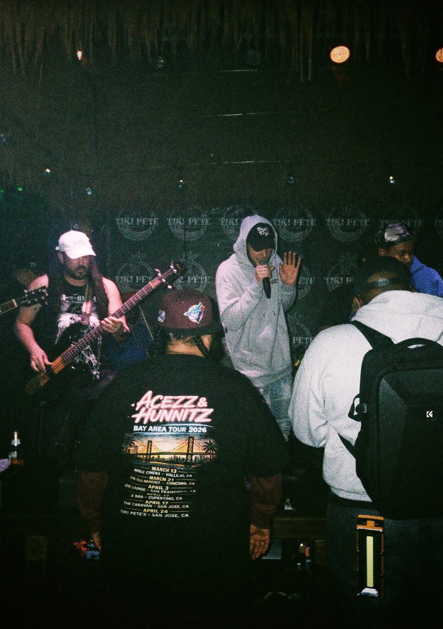 Group of people performing on stage, one person singing into a microphone, others playing guitar, dark background, stage lighting.