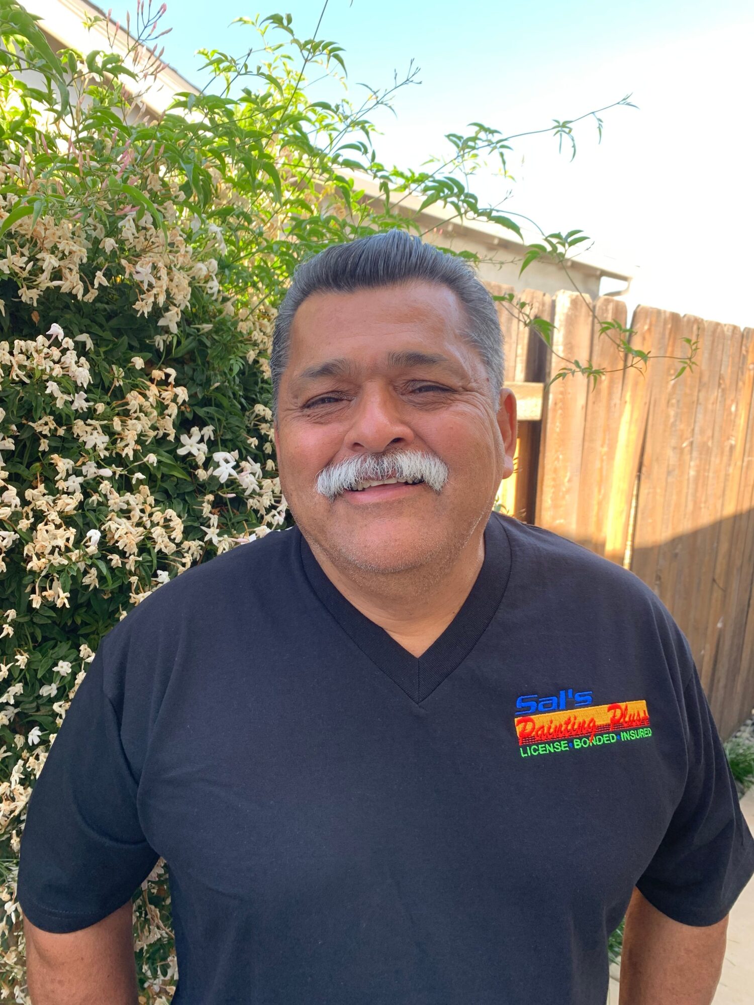 Man with gray hair and mustache smiling outdoors, standing in front of flowering bushes and a wooden fence.