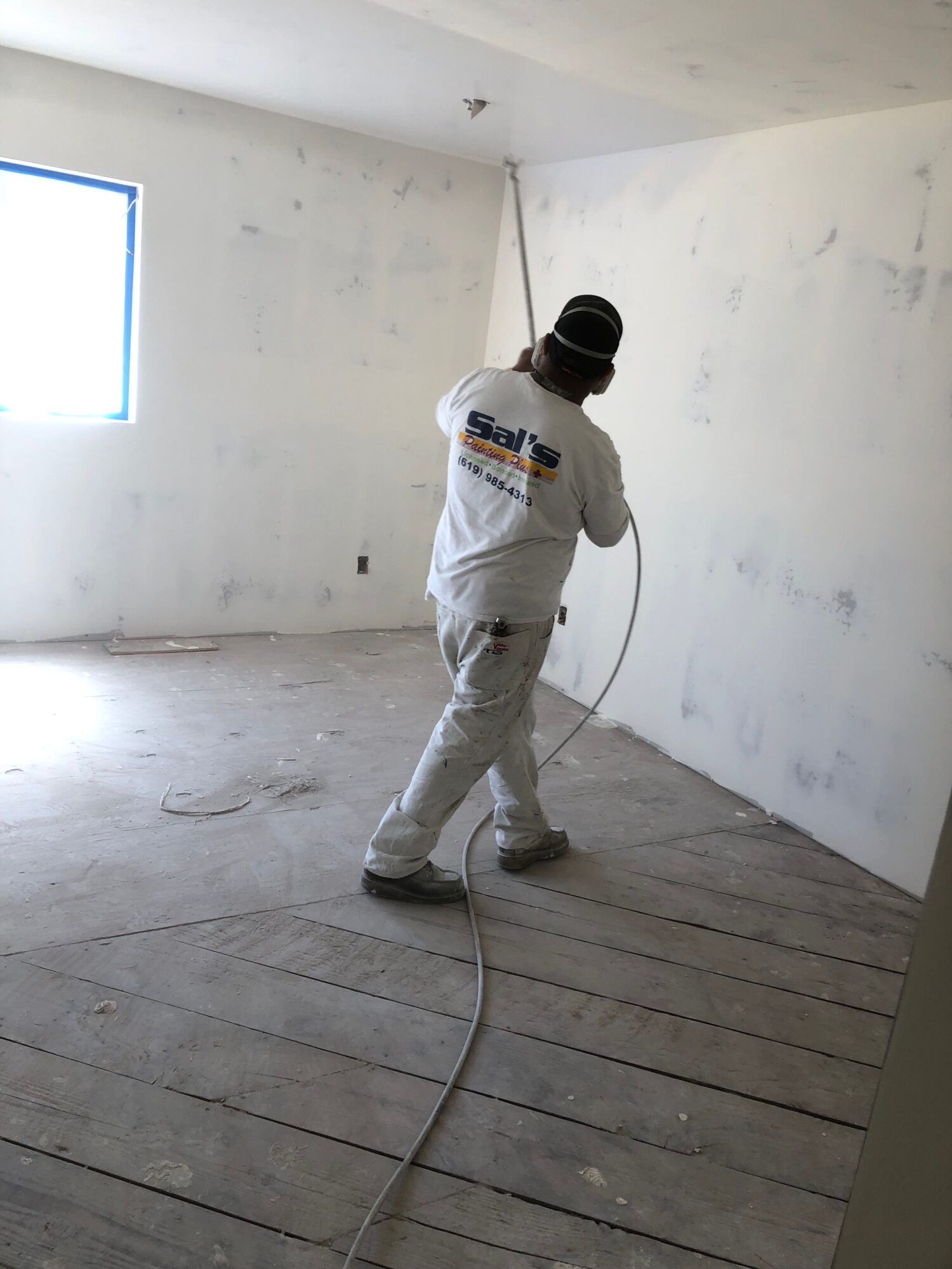 Person in white protective suit and black helmet holding a spray gun in a room with white walls and wooden floor.