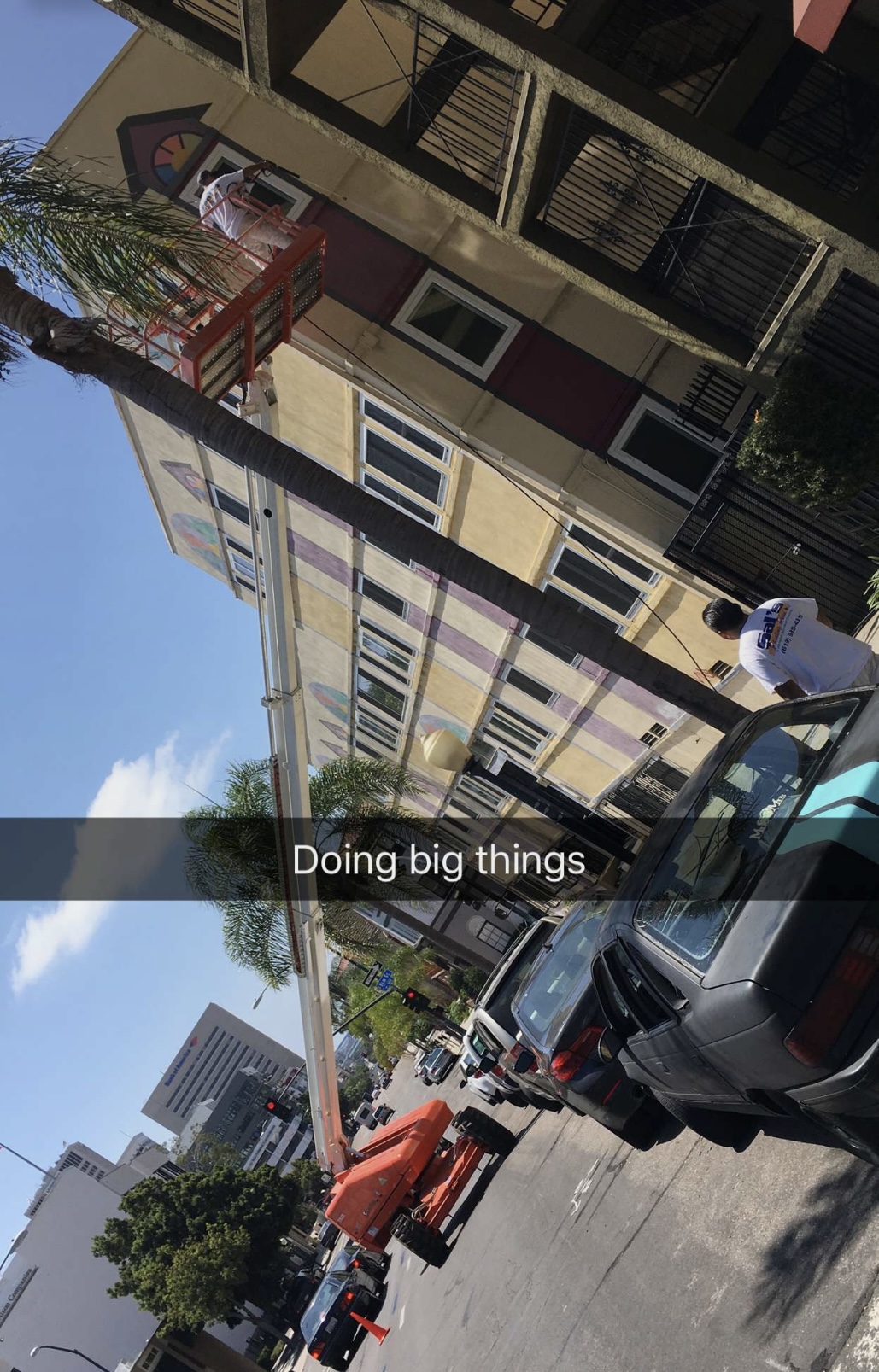 Street view with buildings, parked cars, and people walking, under a clear blue sky.