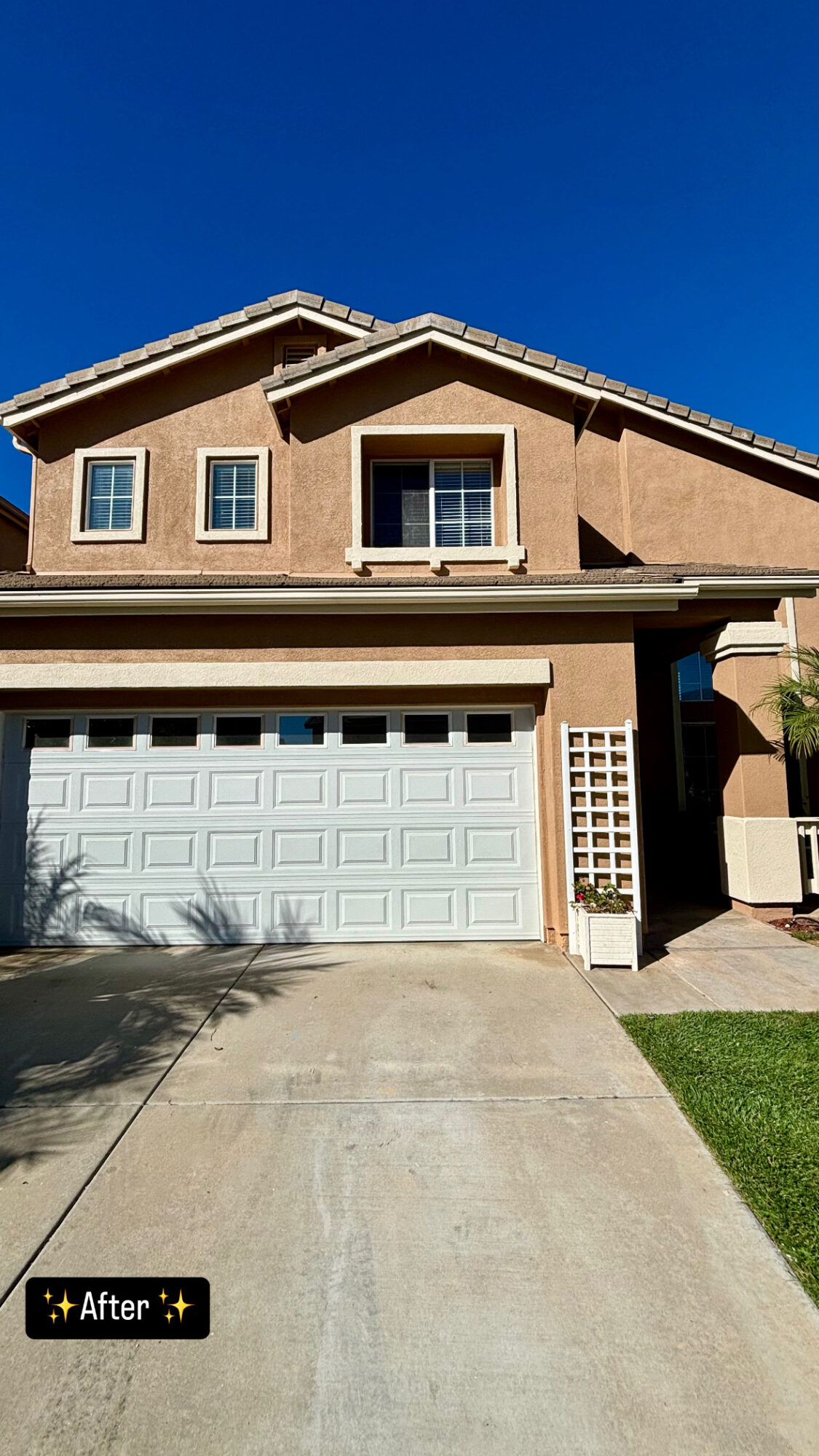 Two-story house with a beige exterior, white garage door, and a small front yard under a clear blue sky.