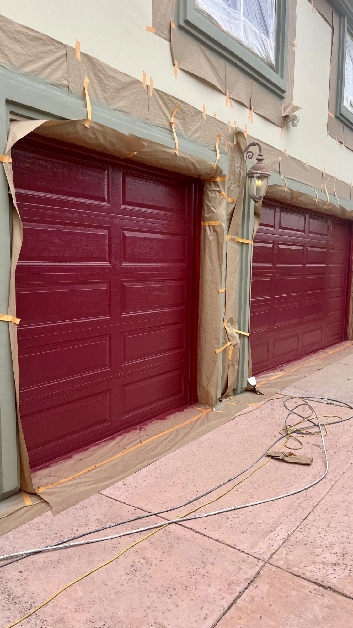 Two closed maroon garage doors with protective wrapping and tape, in front of a building with windows, on a tiled sidewalk.