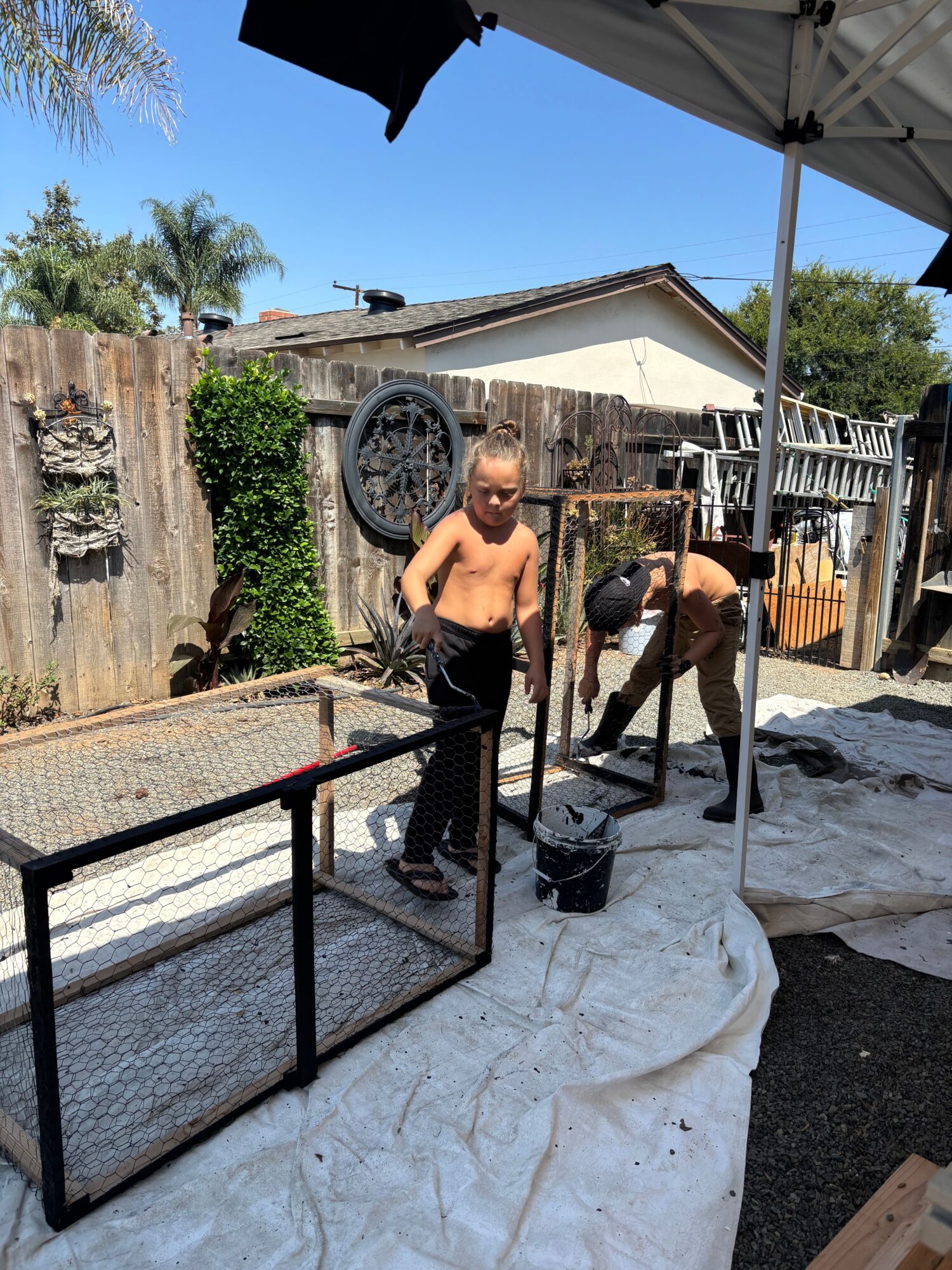 Two shirtless men are outdoors, one sitting on a chair and the other standing nearby, with a backyard and trees in background.