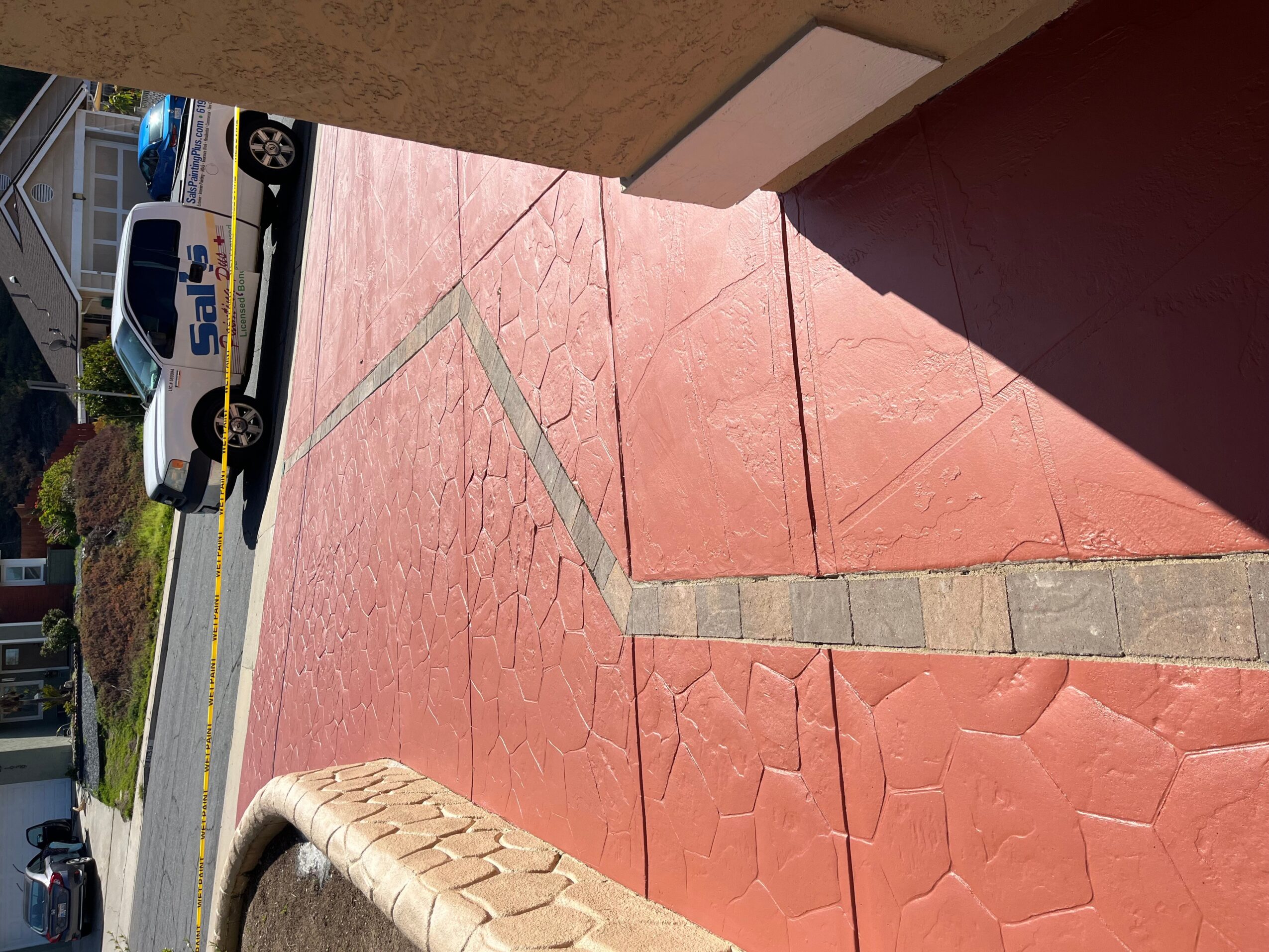 Red sidewalk with gray decorative lines, part of a building wall, and a shadow cast on the ground.