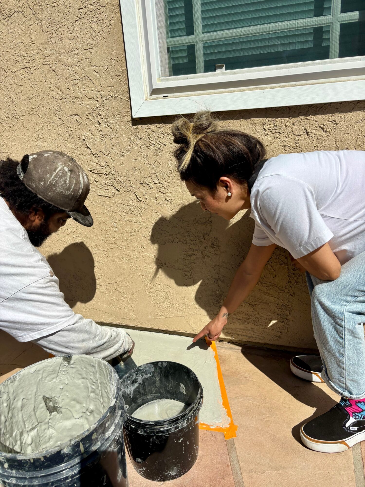 Two people working on a wall, one with a hat and the other with a bun, applying a substance with a trowel.