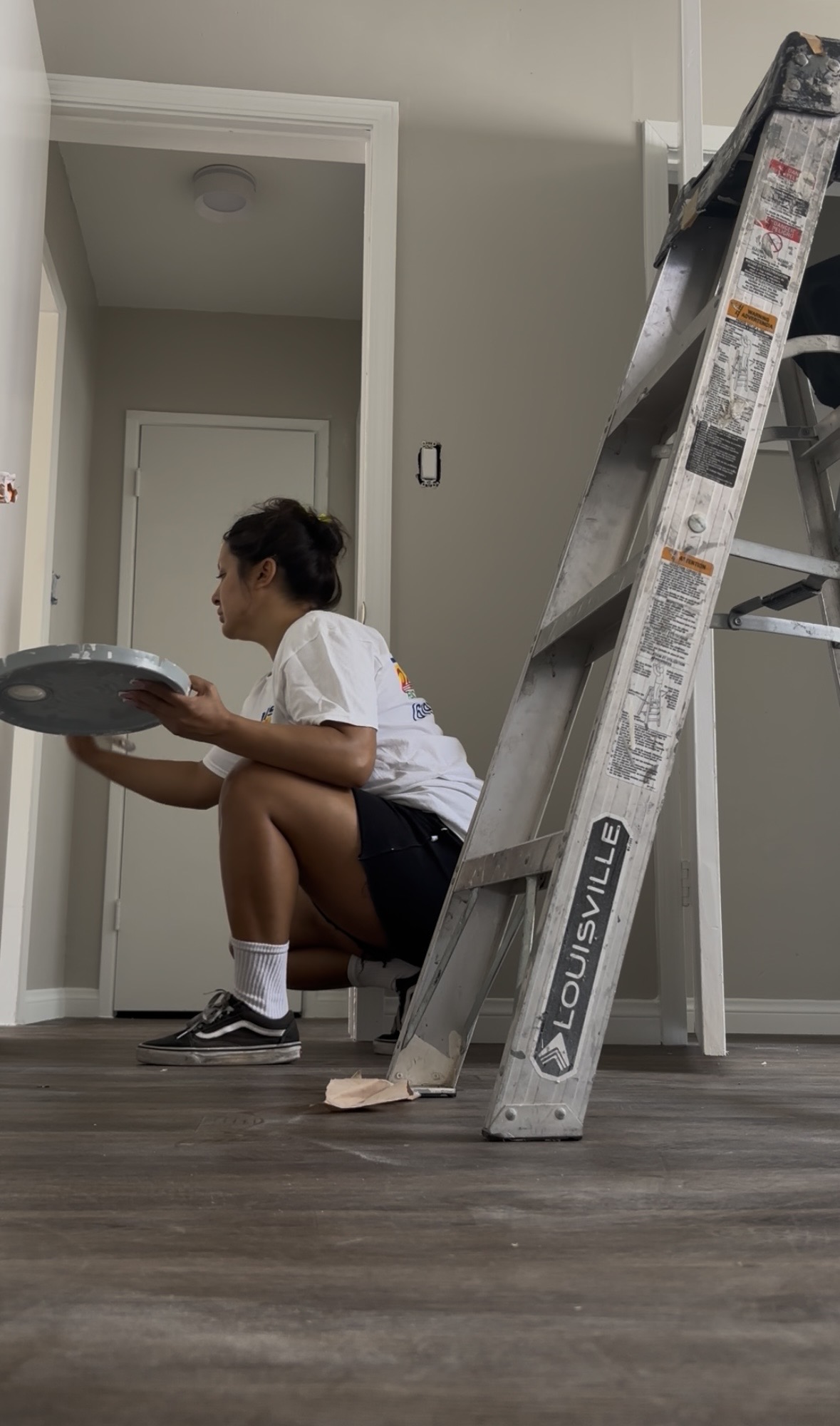Child crouching with a paint palette, ladder nearby, in a room with a door and plain walls.