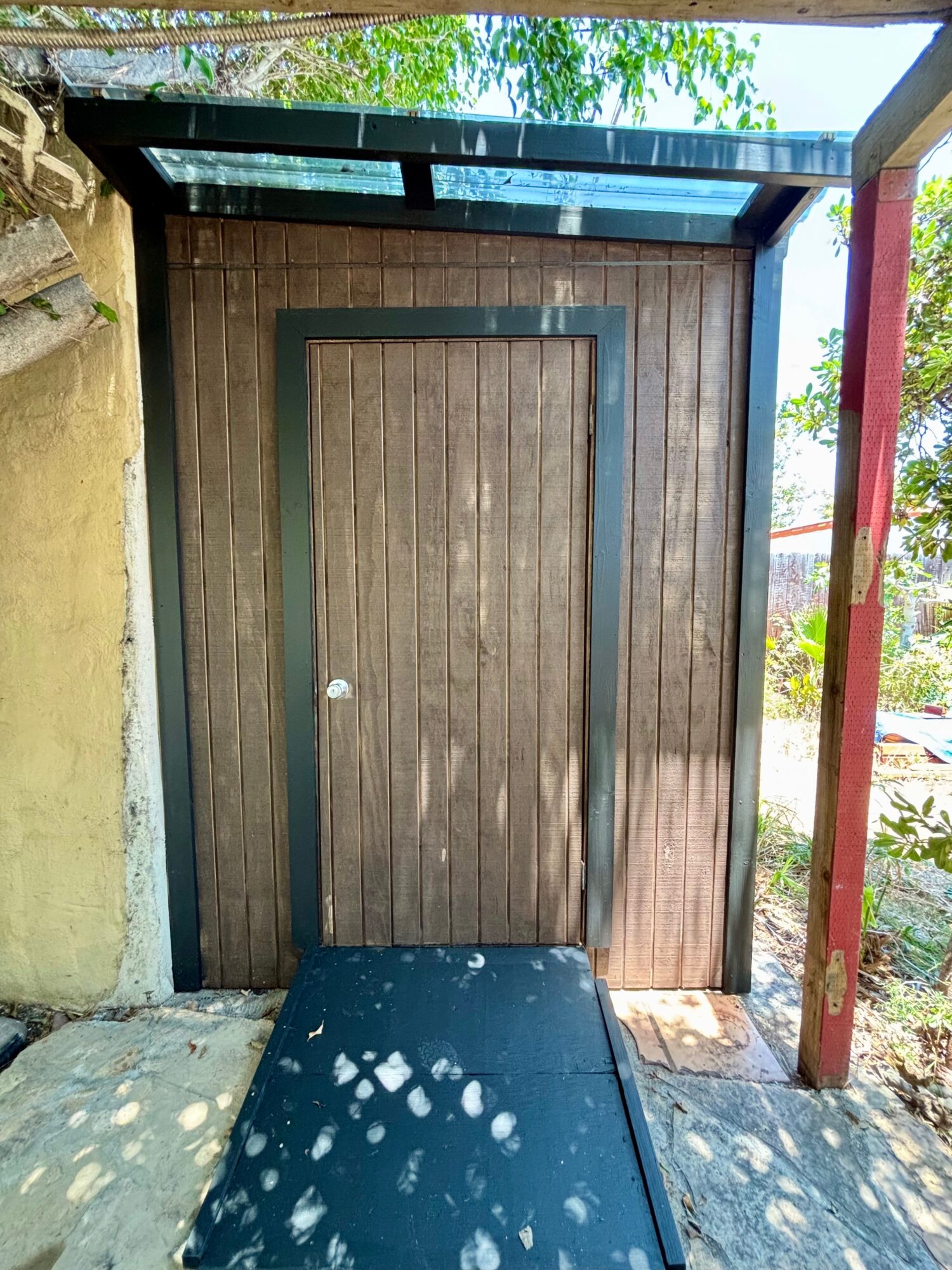 Wooden door with a black frame, set in a wooden wall, outside under a metal roof, with sunlight and shadows.