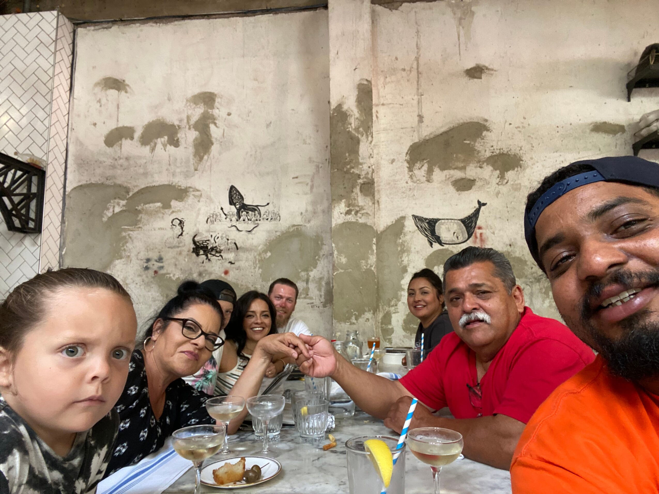Group of people sitting at a table, holding hands, with a wall and butterfly mural in the background.