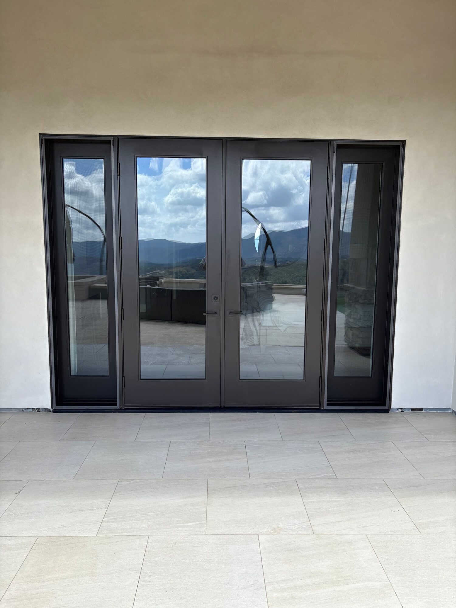 Glass door with black frame, reflecting sky and clouds, leading to an outdoor area, with a tiled floor outside.