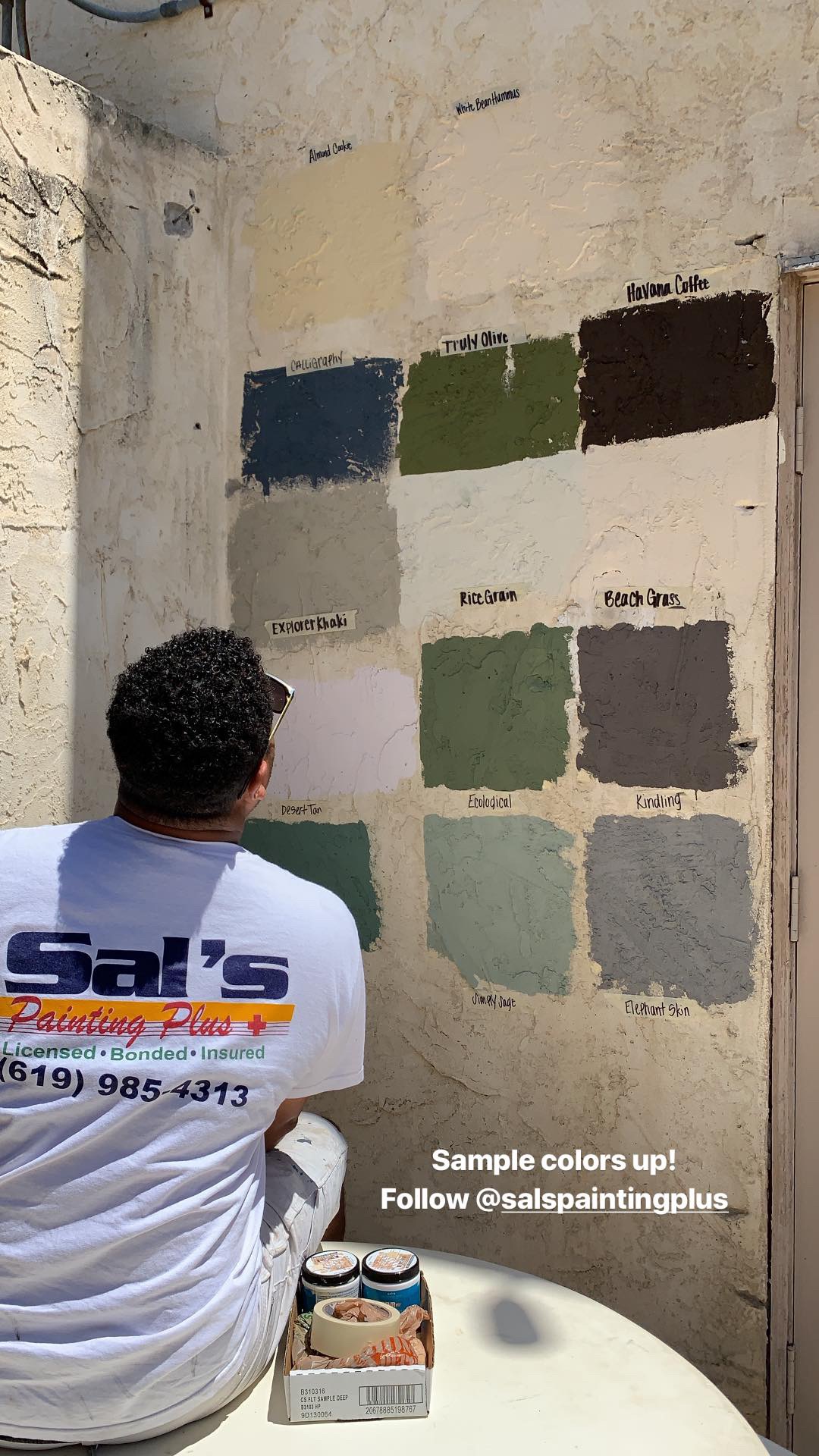 Person with curly hair sitting at a table, painting color swatches on a wall, with paint cans nearby.