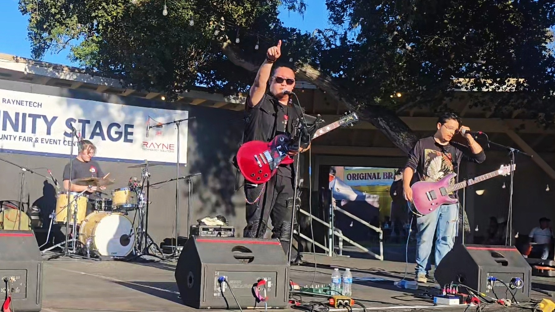 Band performing on stage outdoors with trees in background, three musicians playing instruments, one singing, sunlight, and equipment visible.