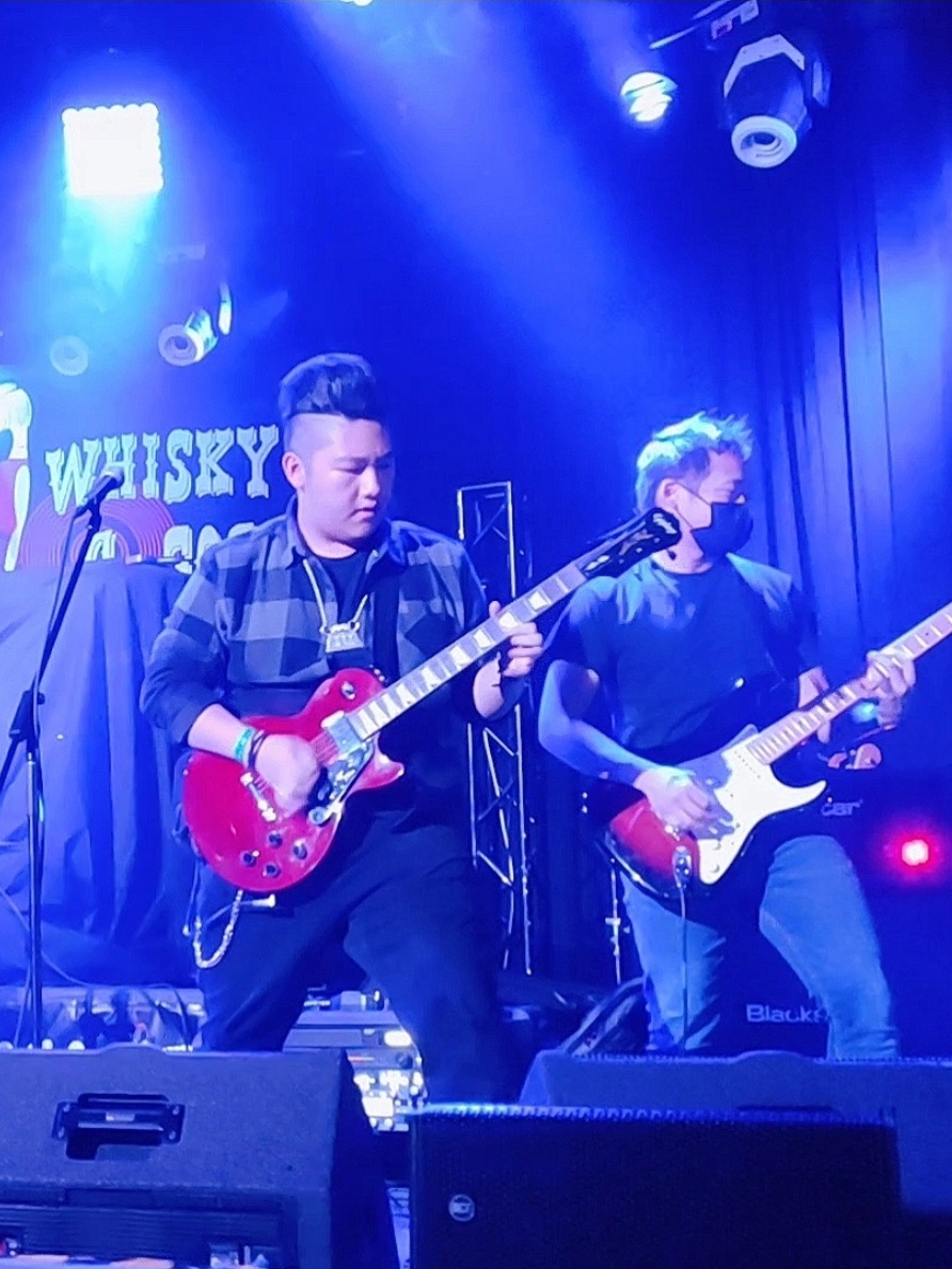Two musicians playing electric guitars on stage with blue lighting and a sign in the background.