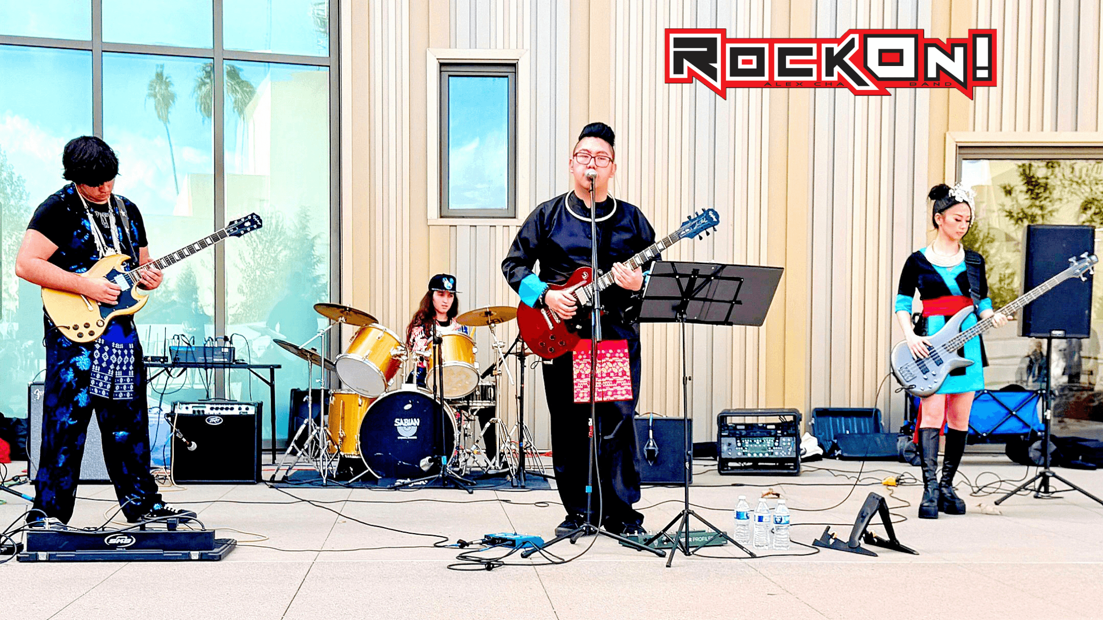 Band performing on stage with four members playing guitars and drums, in a bright indoor setting.