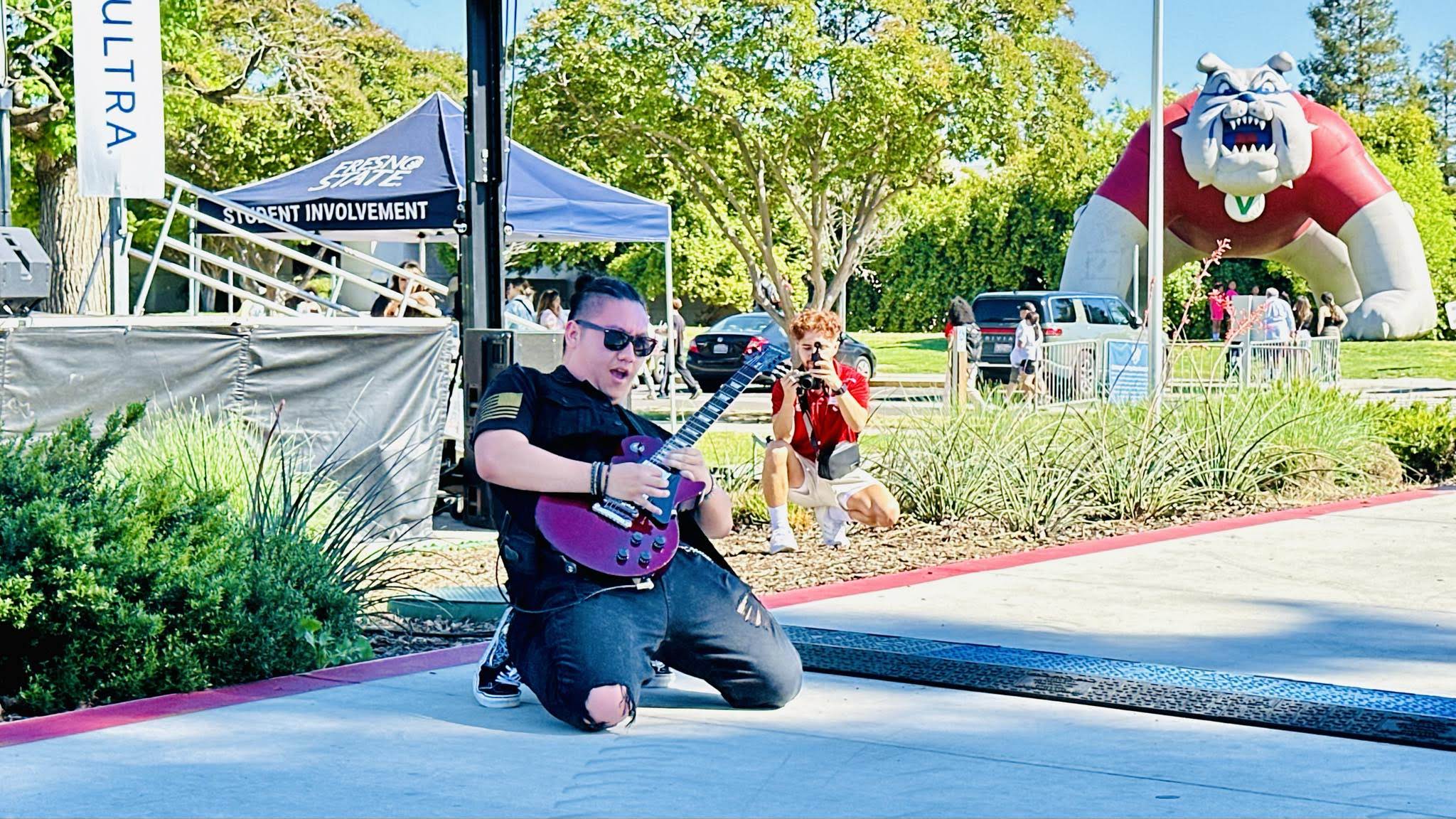 Person kneeling on ground playing purple guitar outdoors with trees and a large dragon sculpture in background.