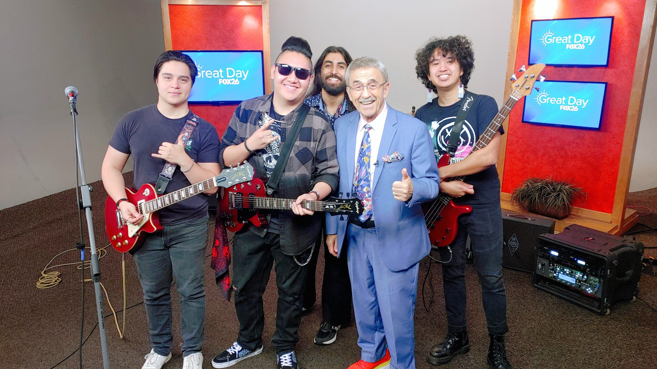 Group of five people, some holding guitars, posing in a TV studio with screens and equipment behind them.