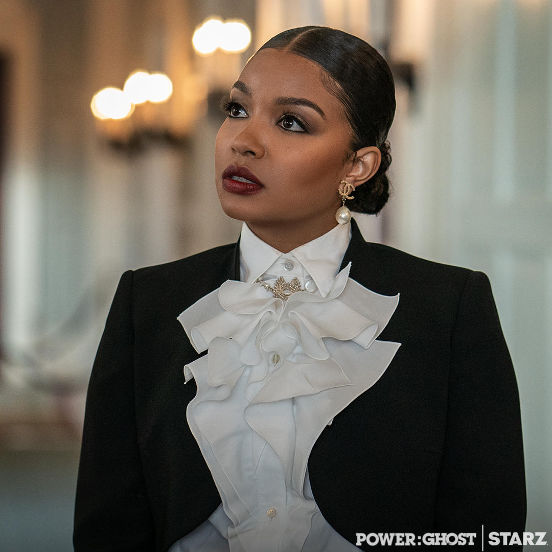 Woman with dark hair in a bun, wearing a black blazer and white ruffled blouse, looking to the side in a formal setting.