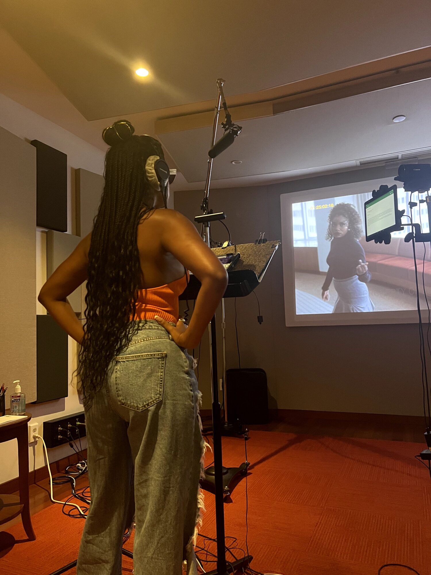 Woman with long hair stands in a recording studio, facing a screen showing a person, with equipment around her.