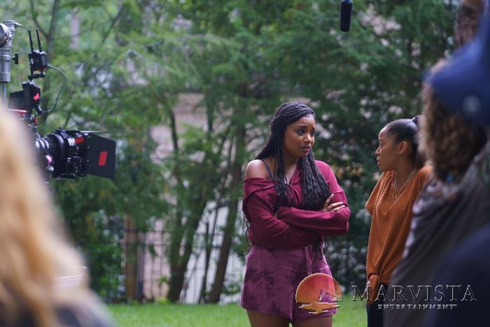 Two women engaged in conversation outdoors with trees in the background, surrounded by a film crew and equipment.