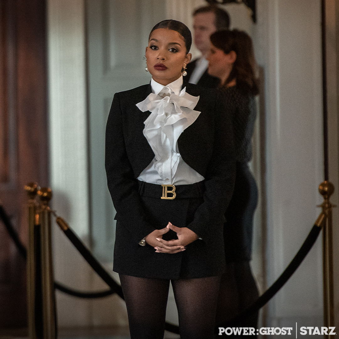 Woman in formal black suit with white ruffled blouse, standing with hands clasped, in a room with people in the background.